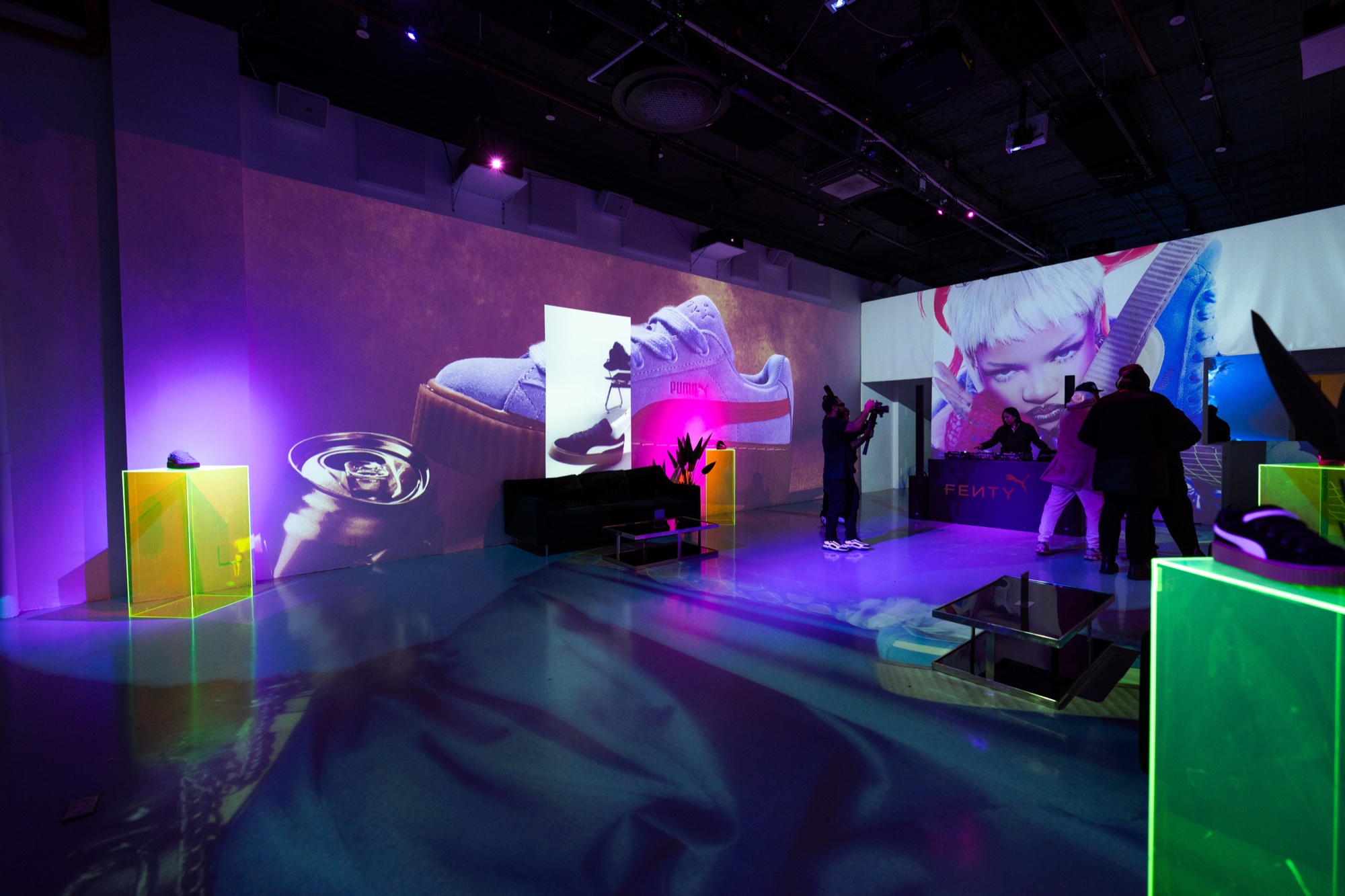 Wide view of a projection filled room with green neon plinths, a sofa and a pink haired mural