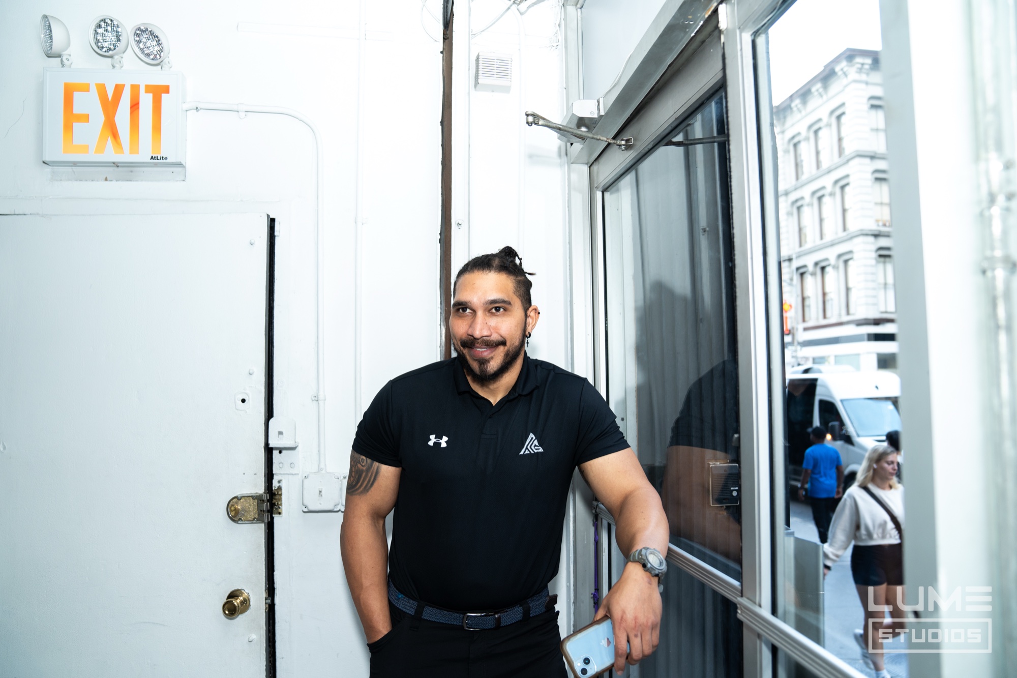 Man in a black polo with a topknot leans in a white doorway under an exit sign, holding a phone