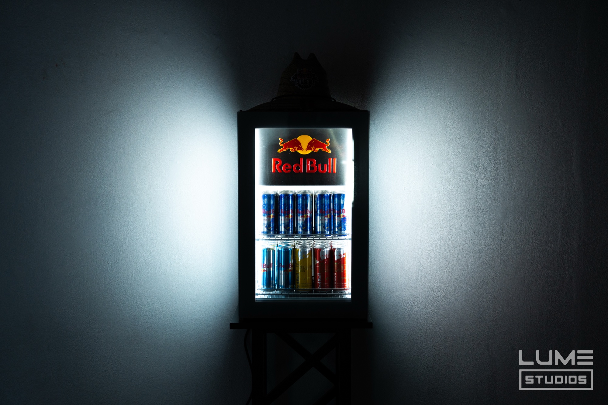 Lit Red Bull mini fridge stocked with cans glowing on a dark grey wall
