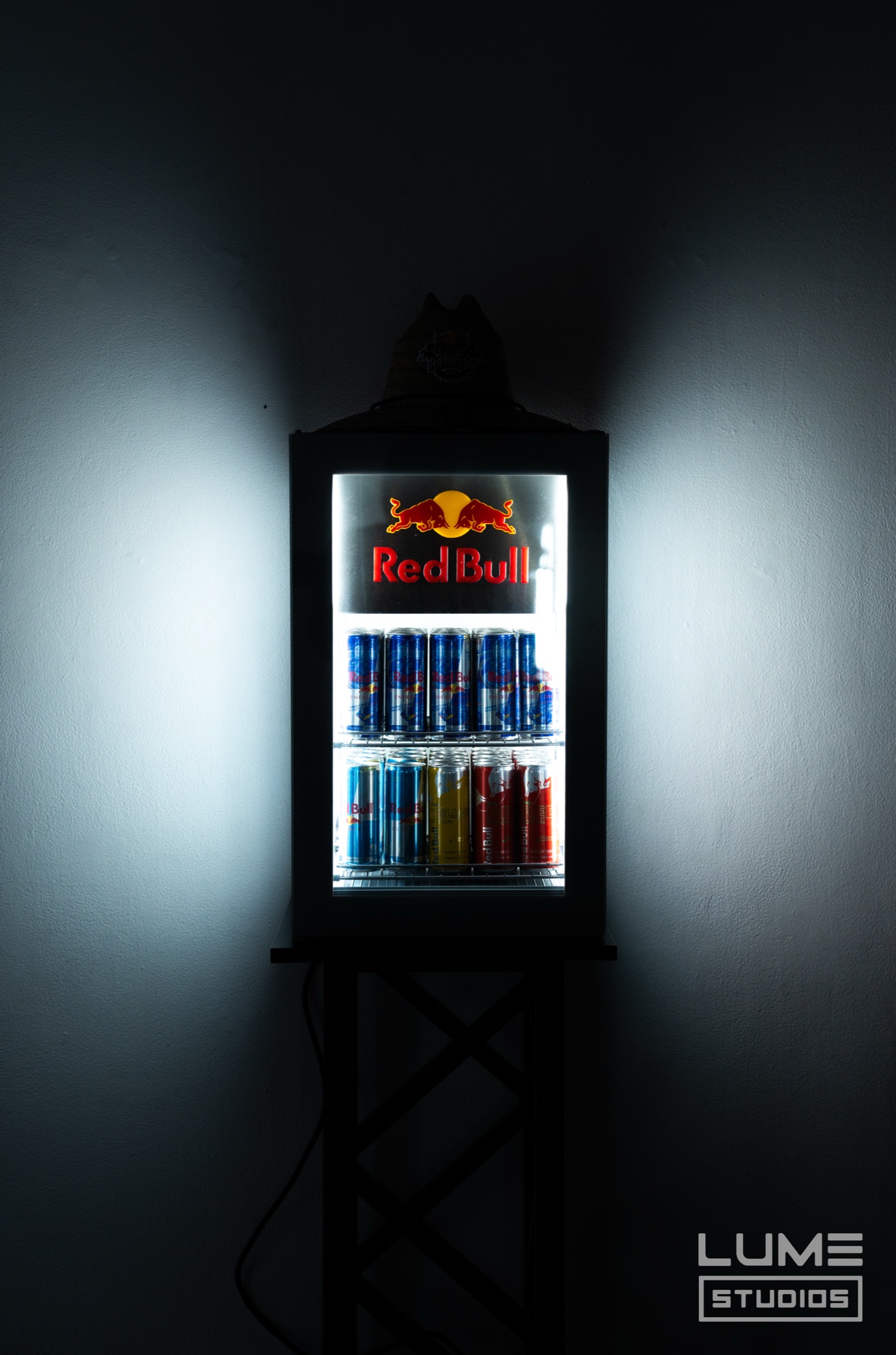 Small glowing Red Bull fridge stocked with cans mounted on a dark wall with light spilling either side