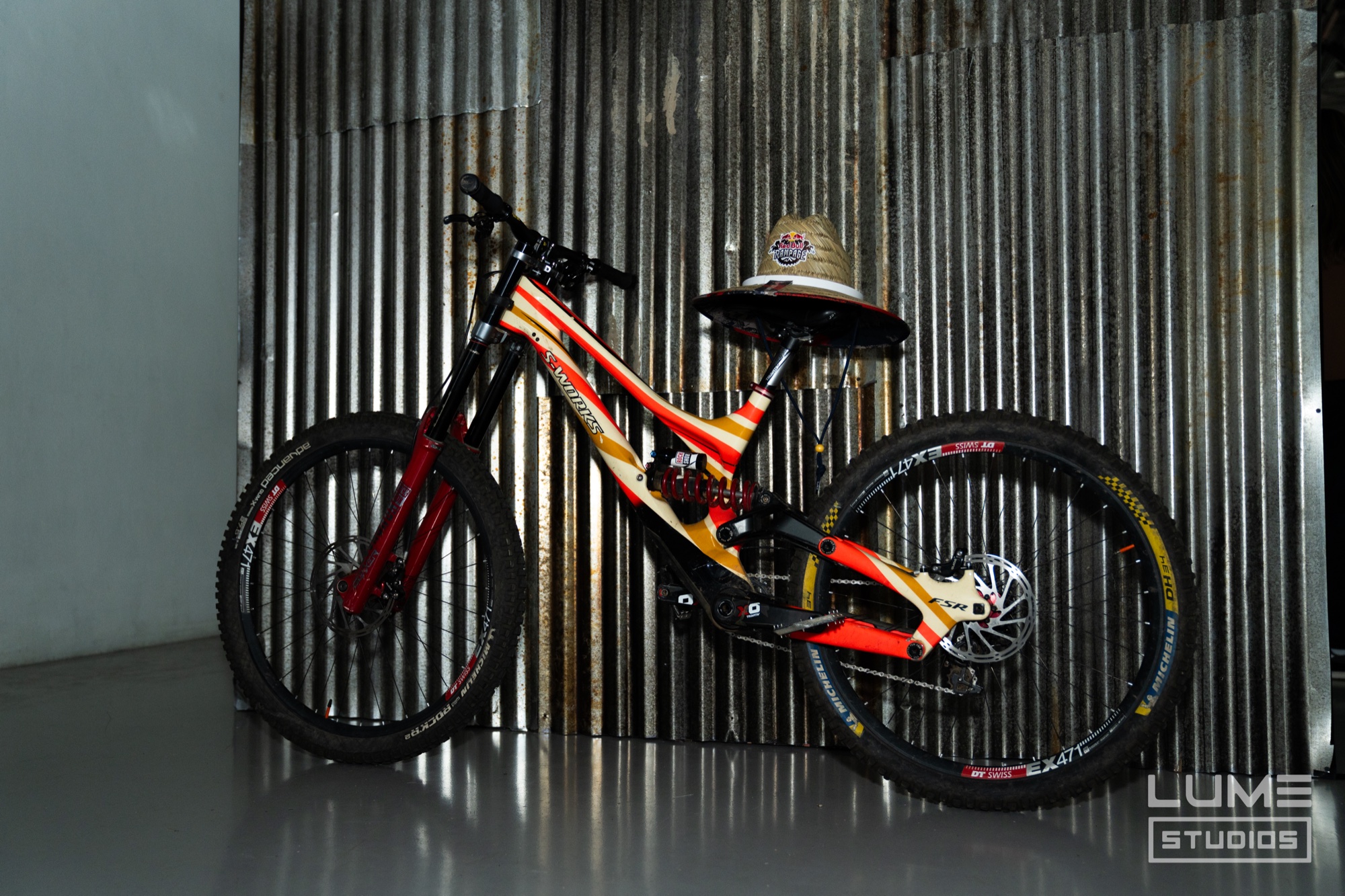 Red and yellow downhill mountain bike with a straw hat on its seat against a corrugated metal wall