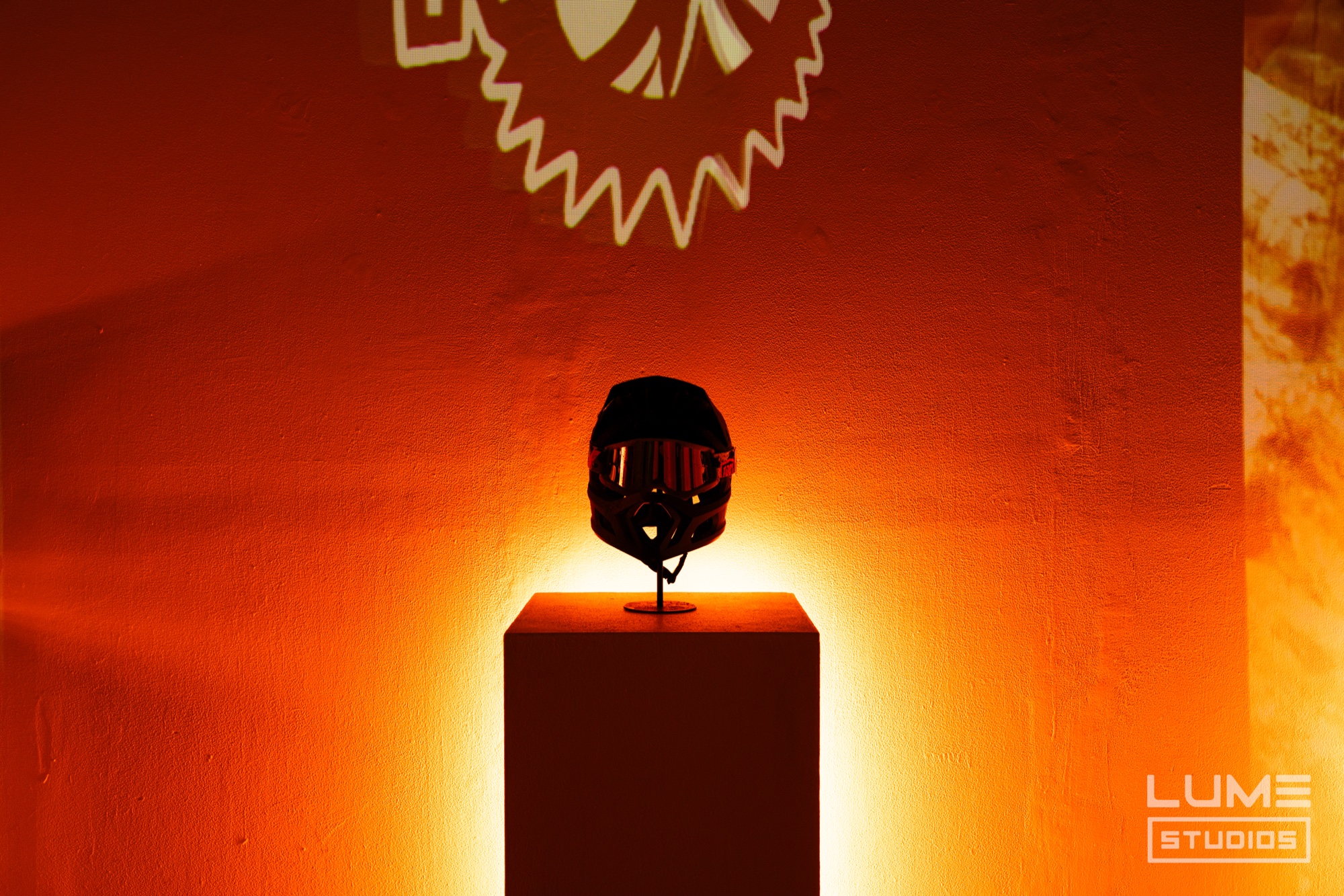 Black full face helmet on a plinth lit from below in orange with a saw blade logo projected above