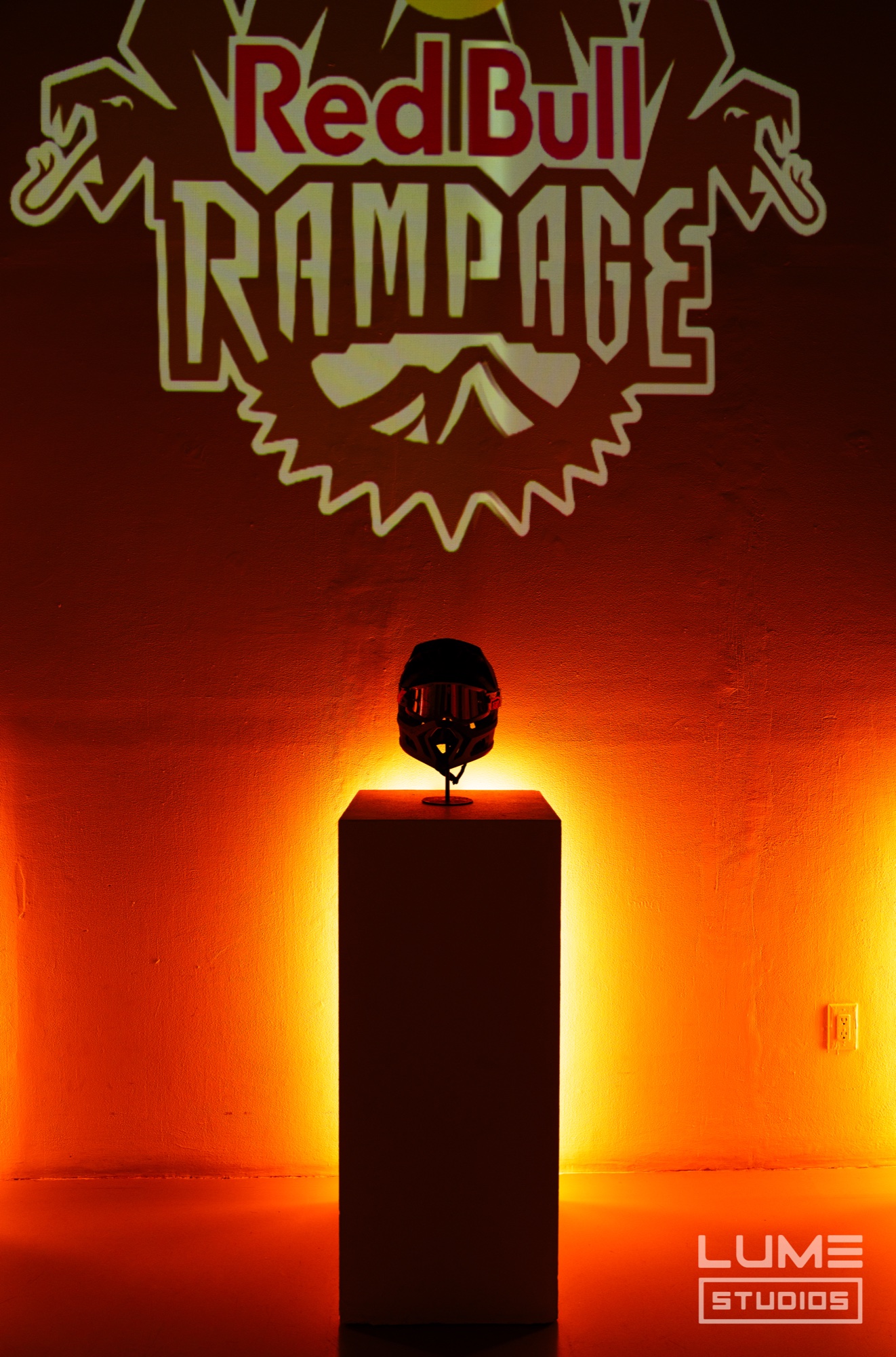 Full face helmet on a white plinth glowing orange beneath a projected Red Bull Rampage logo