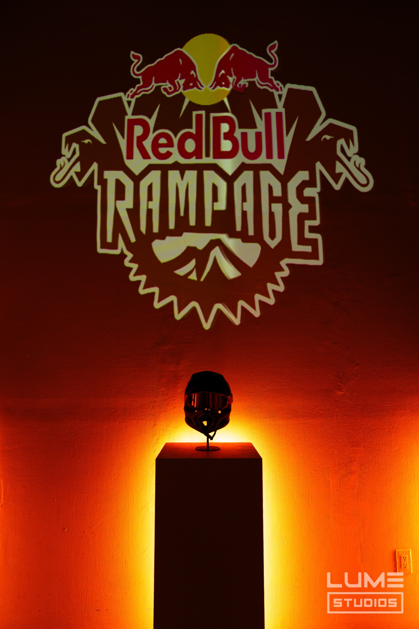 Red Bull Rampage logo projected on an orange lit wall above a bike helmet on a glowing plinth