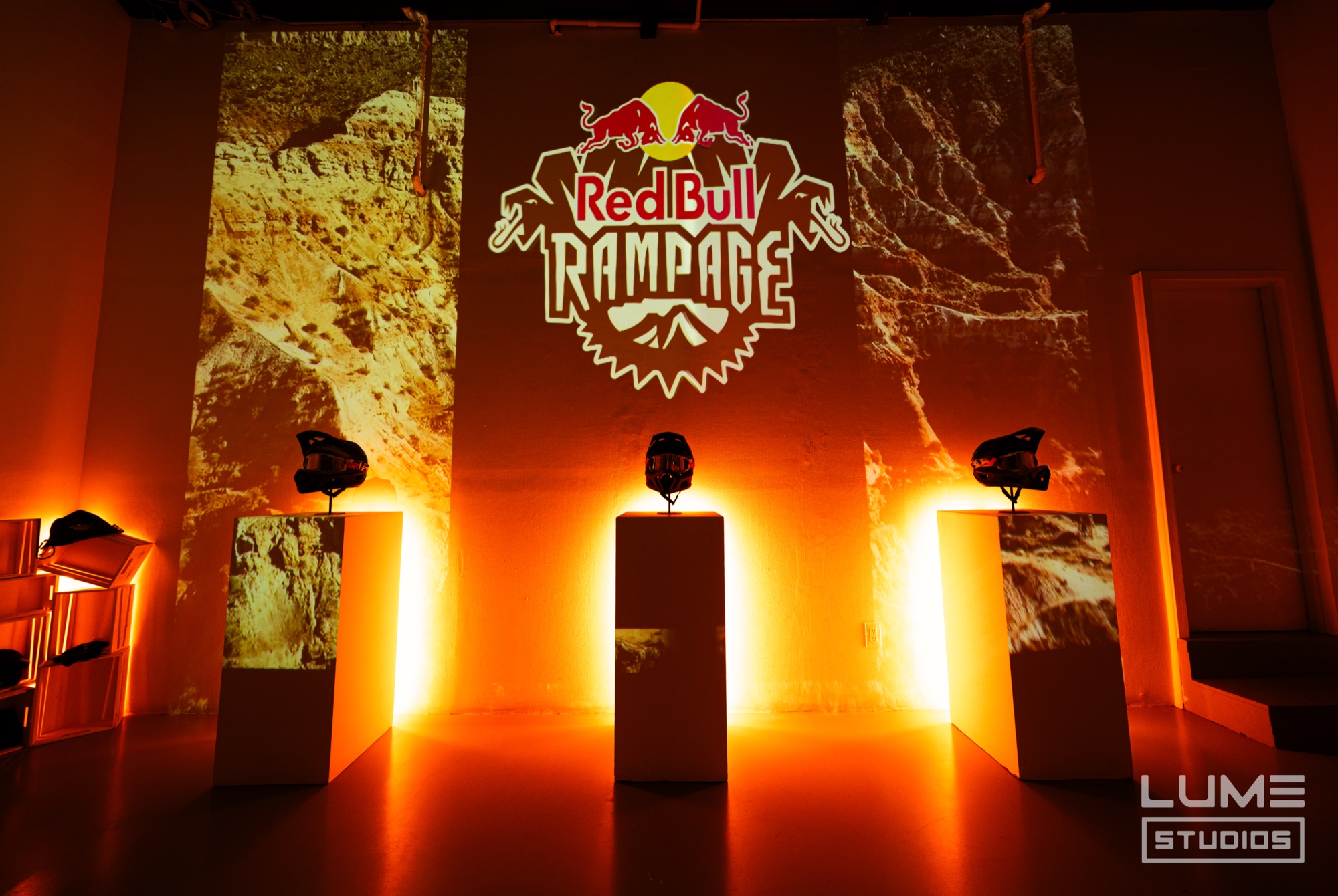 Three bike helmets on glowing plinths under a projected Red Bull Rampage logo and orange canyon rock walls