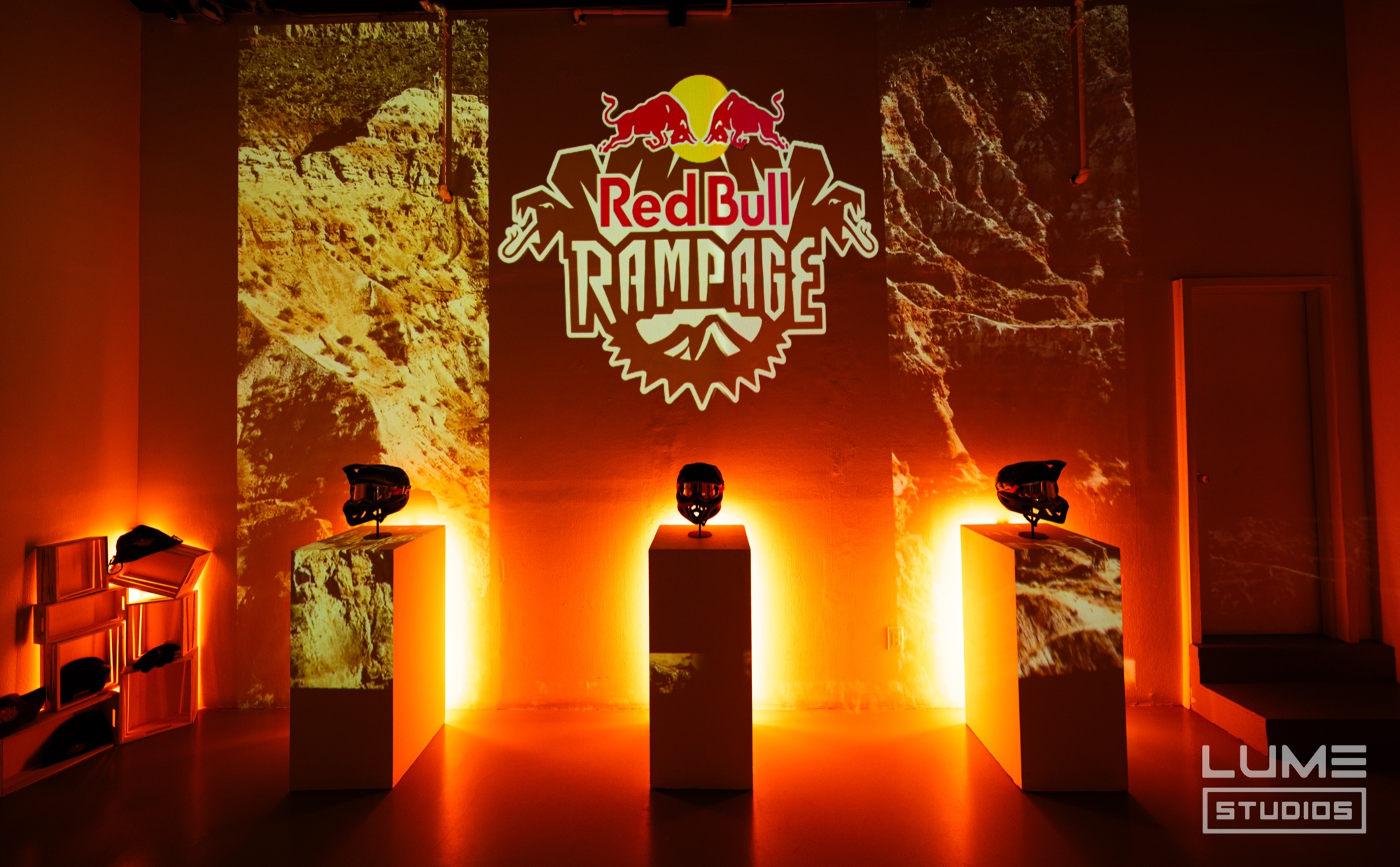 Three helmets on glowing orange plinths before a Red Bull Rampage logo projected over desert cliffs