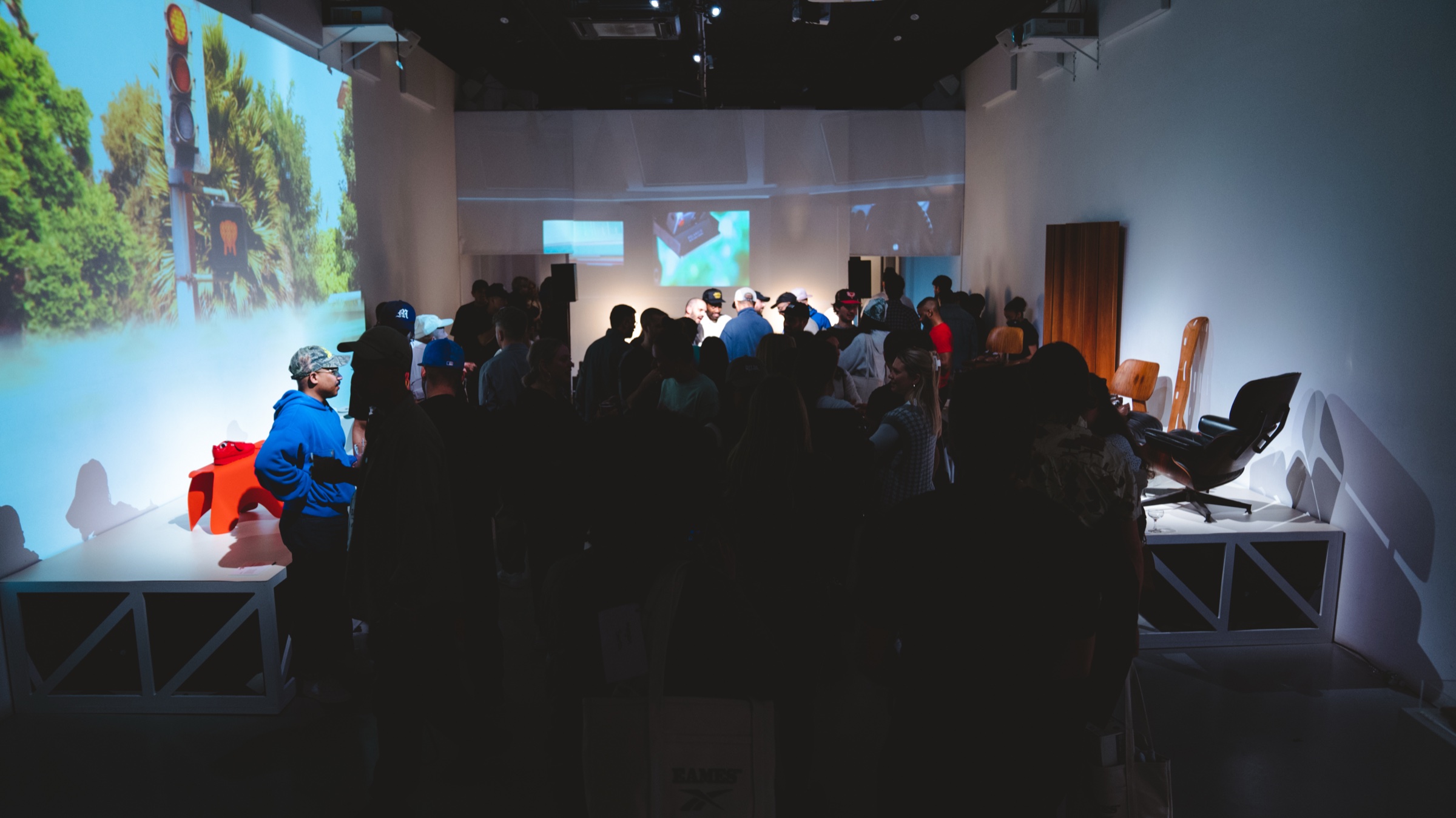 Crowd fills a dark gallery between a projected forest scene and a lounge chair on a platform