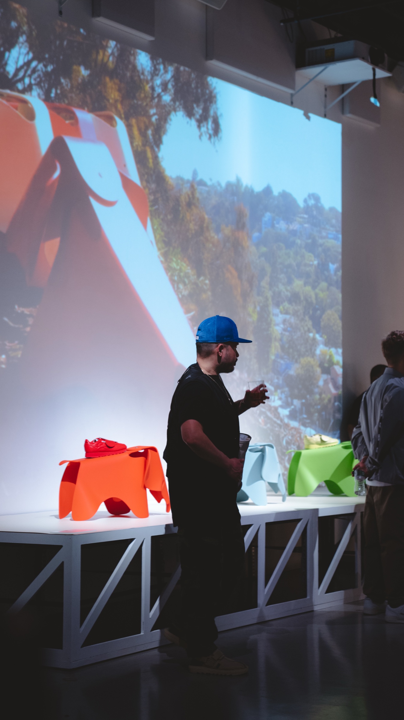 Man in a blue cap holds a drink beside orange, blue and green Eames elephant stools under a projected hillside