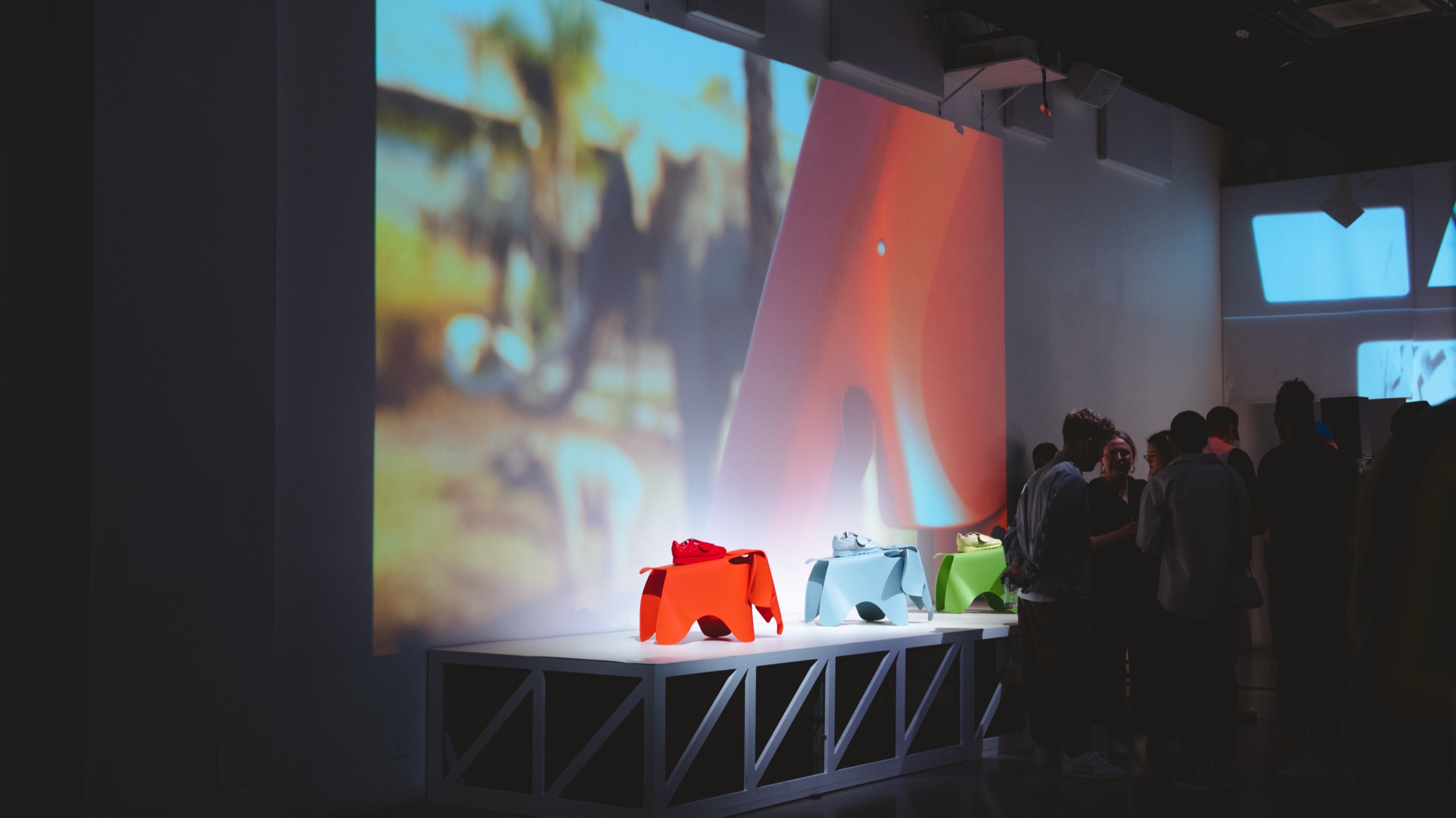 Red, blue and green Eames elephant stools on a white platform beneath a projection of palm trees
