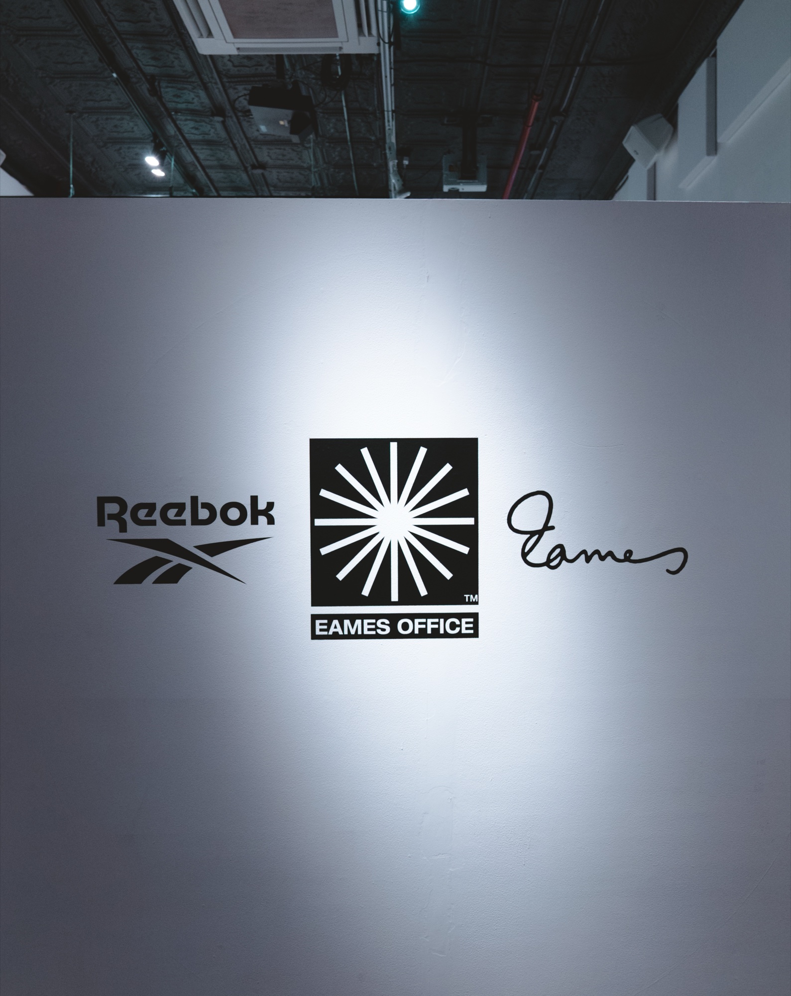 Reebok, Eames Office and Eames signature logos printed on a grey wall under a single spotlight
