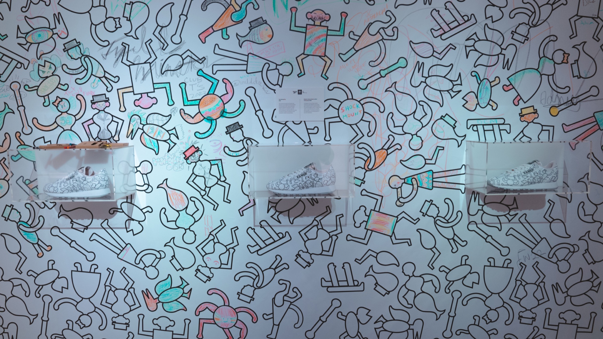 Three sneakers in lit display boxes on a wall covered in colourful line drawn figure patterns