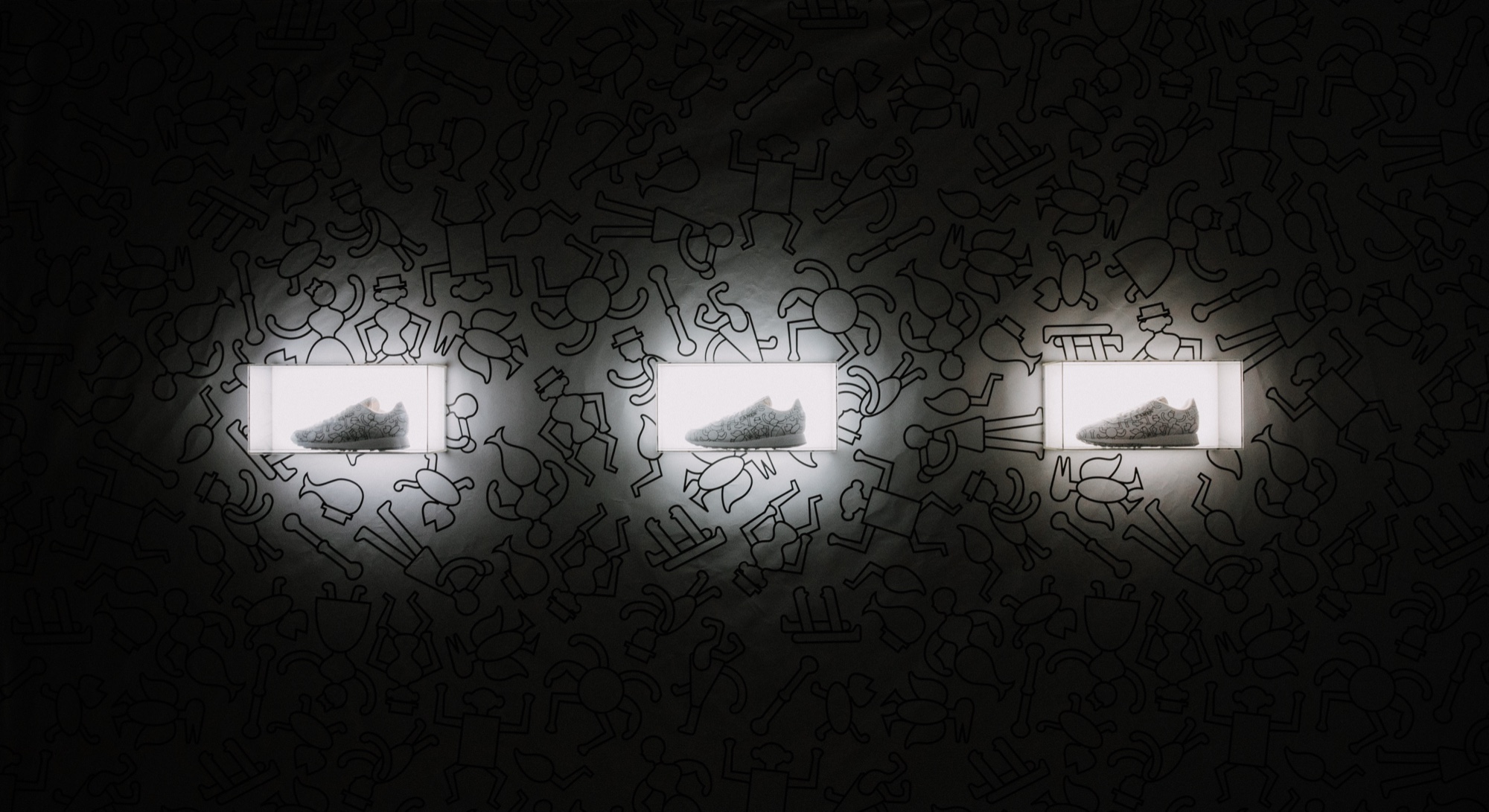 Three backlit boxes each holding a white sneaker set in a dark wall of black line drawings