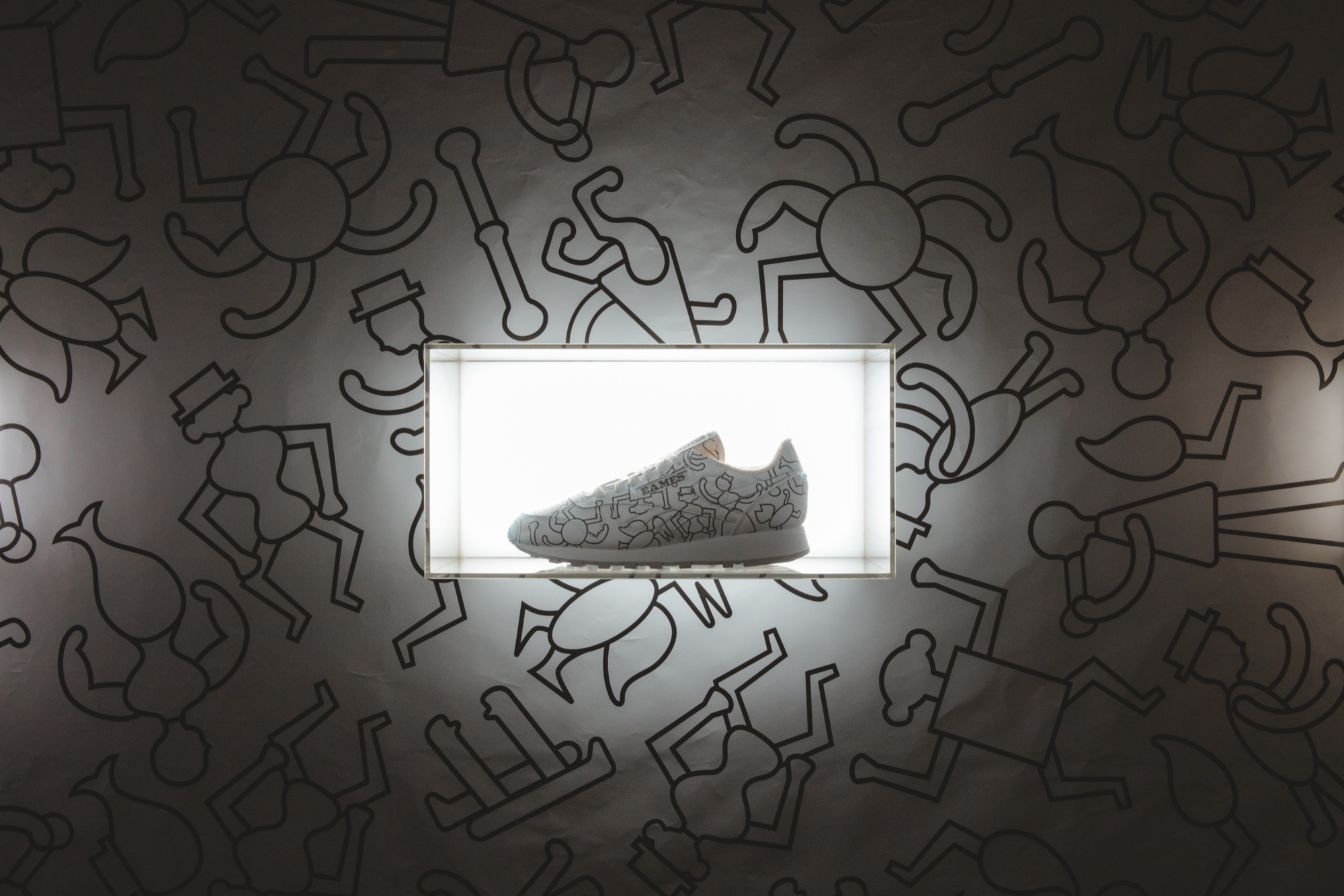White sneaker printed with black line figures glowing in a lit wall box amid a matching wall pattern