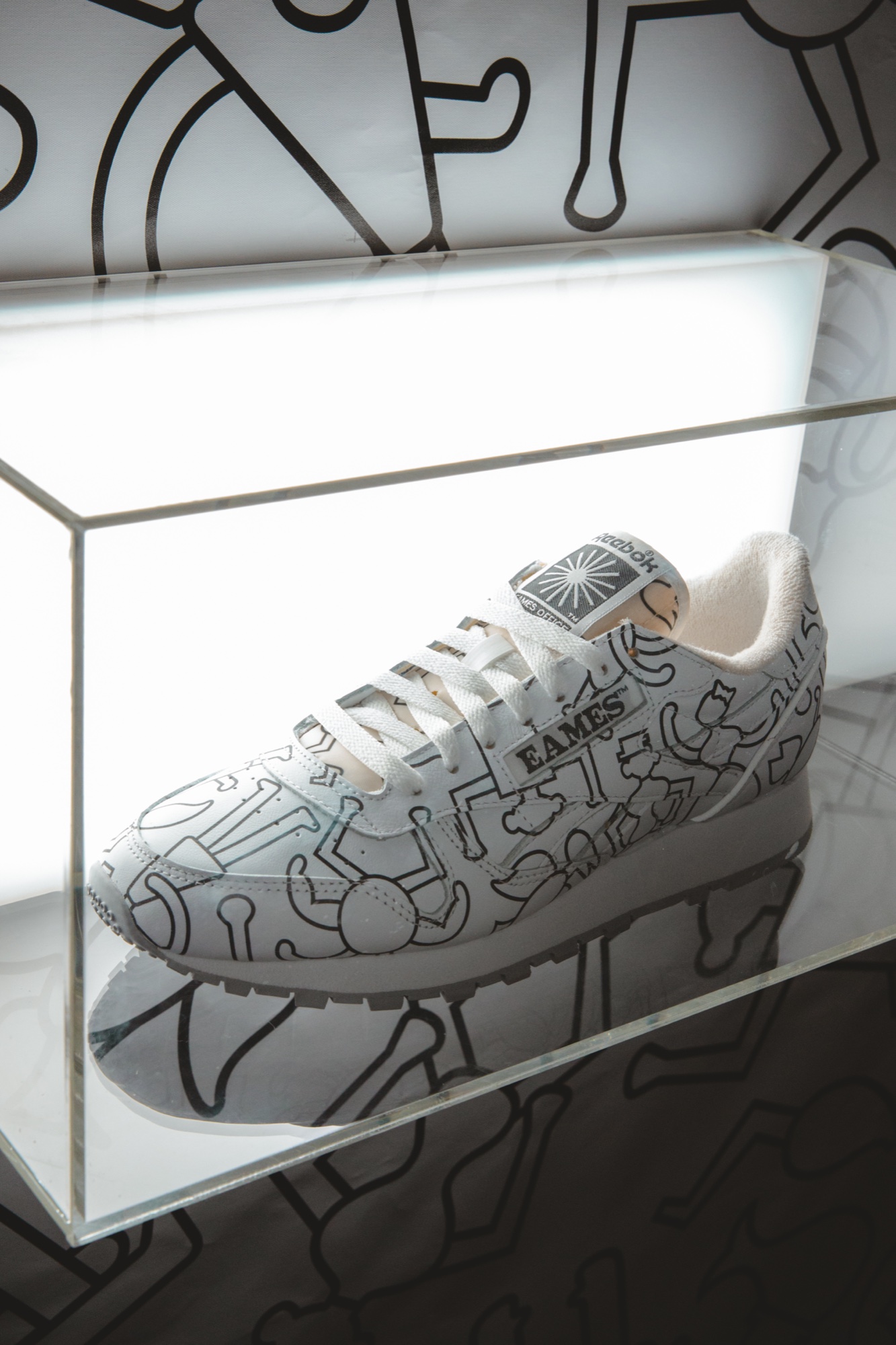White Reebok sneaker covered in black line figures inside a glowing acrylic case, Eames on the side