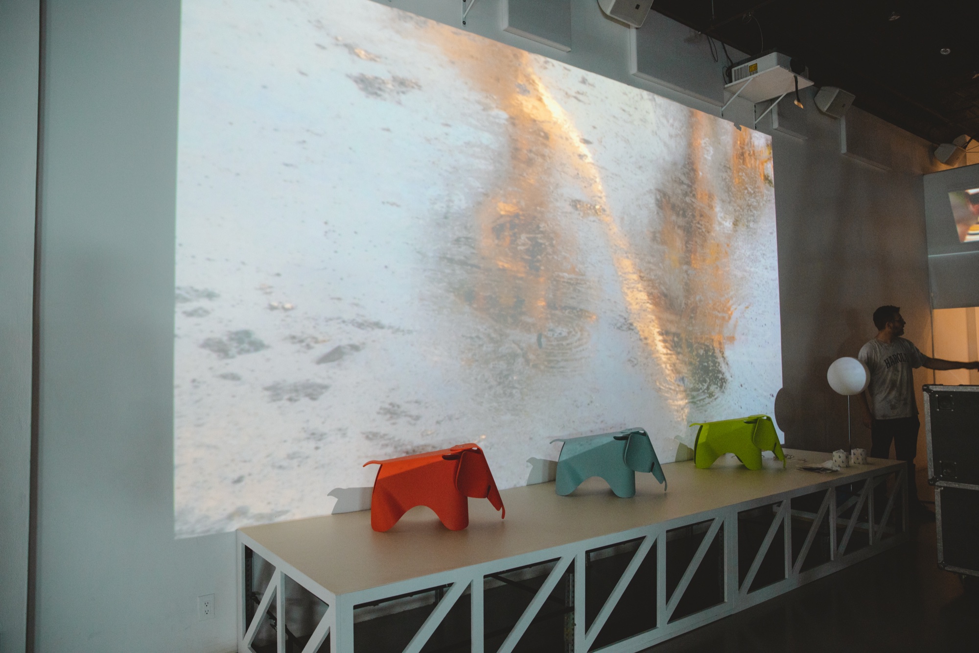 Red, teal and green Eames elephant stools on a white truss platform beneath a snowy mountain projection