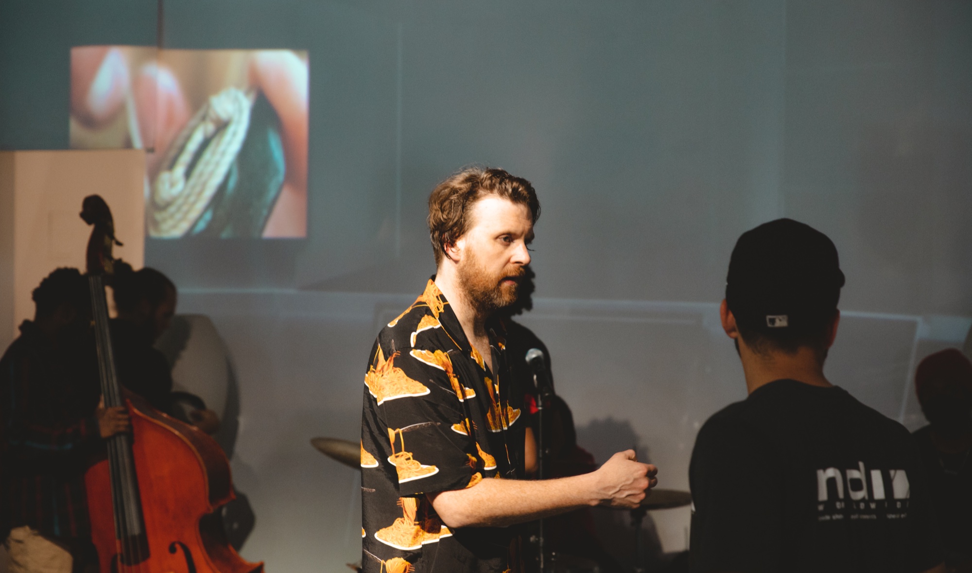 Bearded man in a black and orange print shirt speaks beside a double bass player under a projected close up