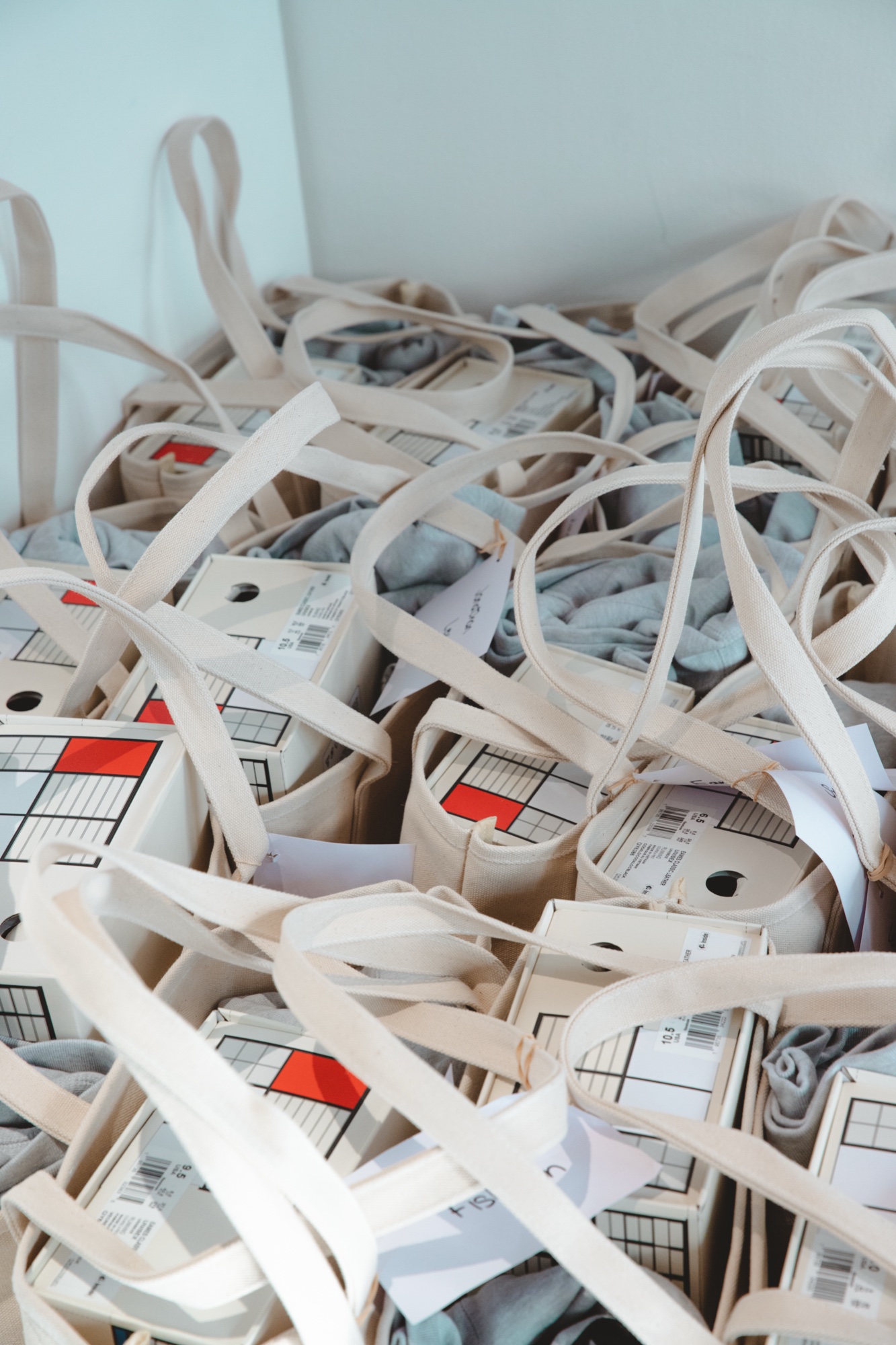 Pile of cream tote bags holding shoe boxes with red, white and black labels and grey tissue