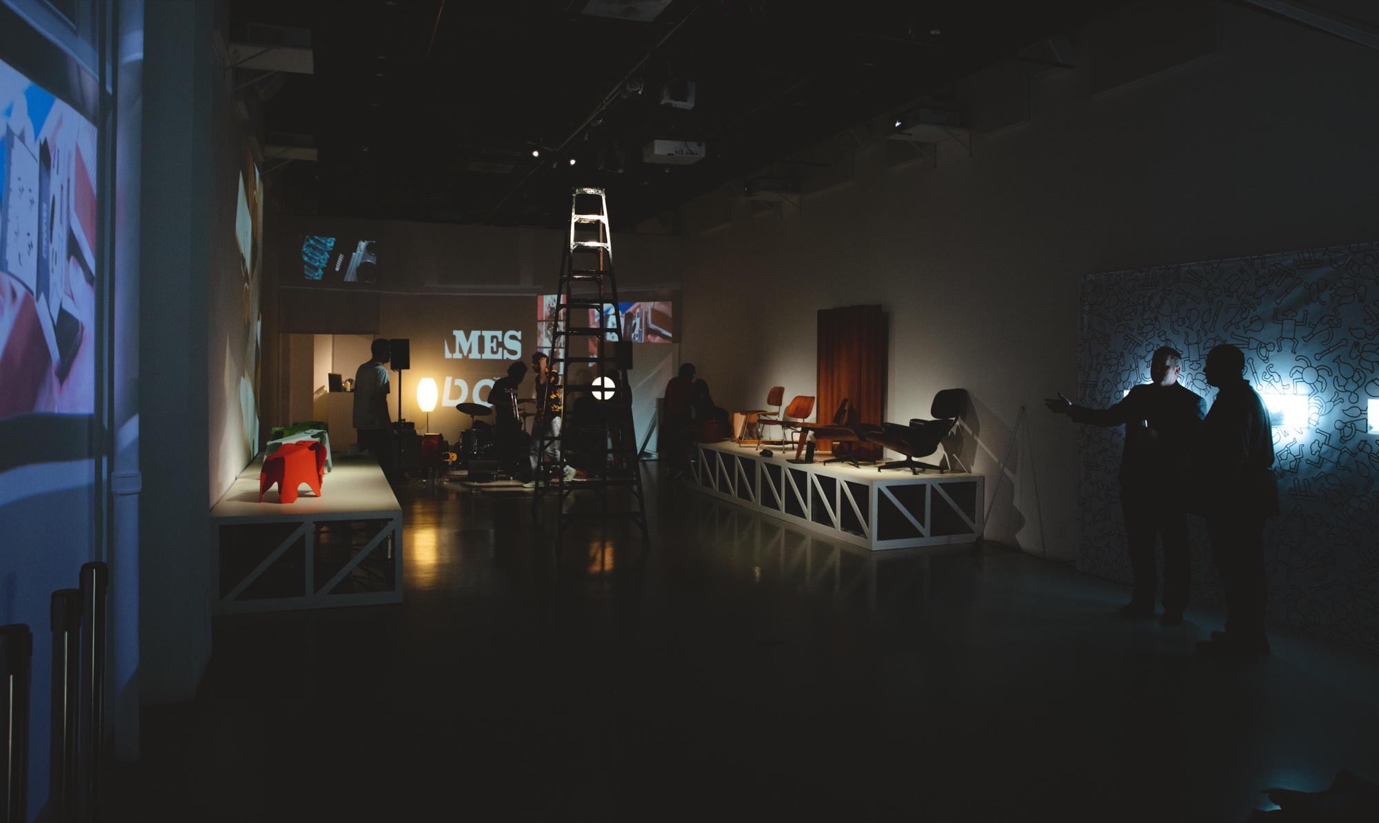 Dim room with a tall ladder, low white platforms, a projection wall and two guests at the right