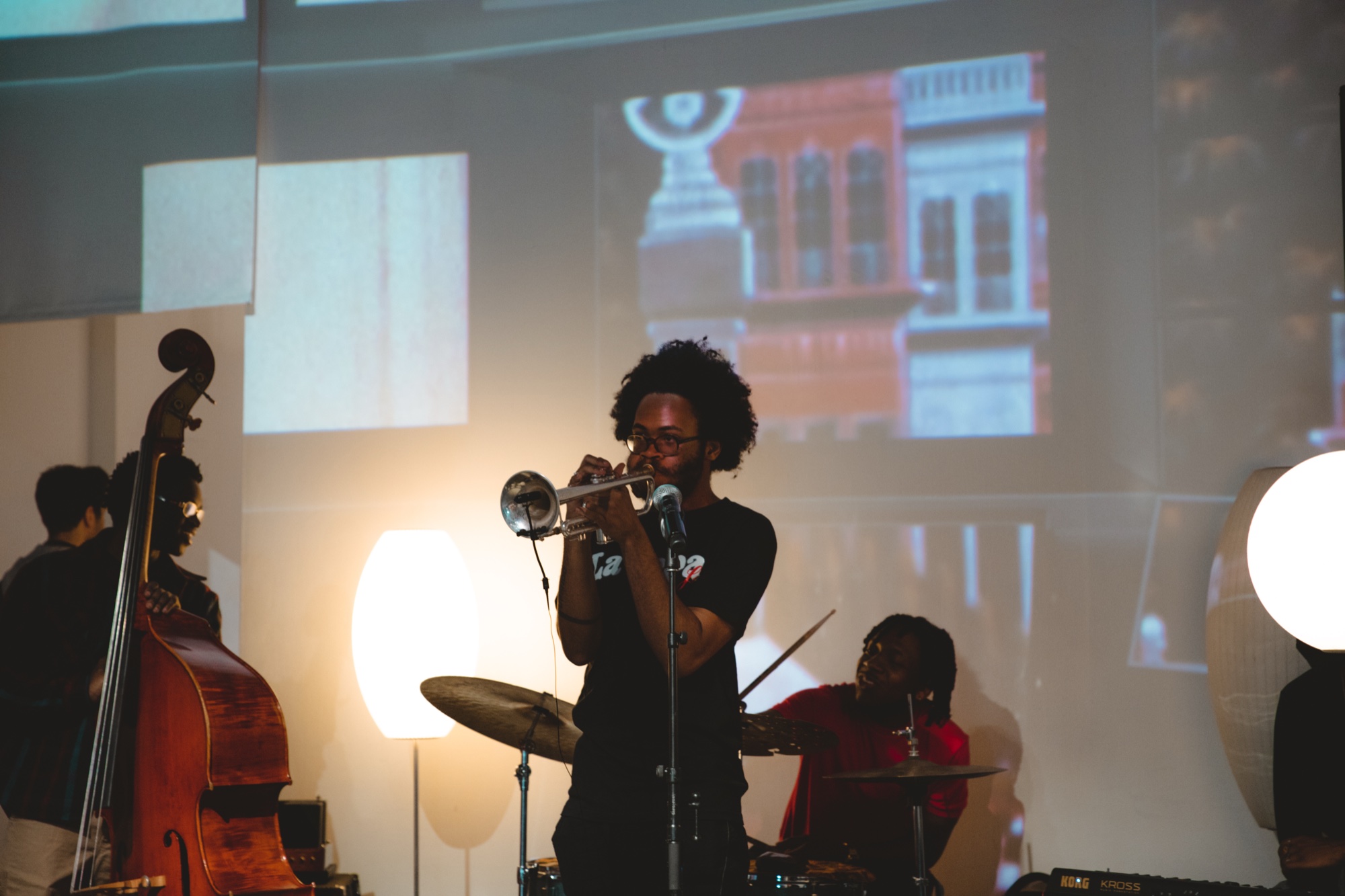 Trumpet player with a large afro performs beside an upright bass and drummer in front of projected video