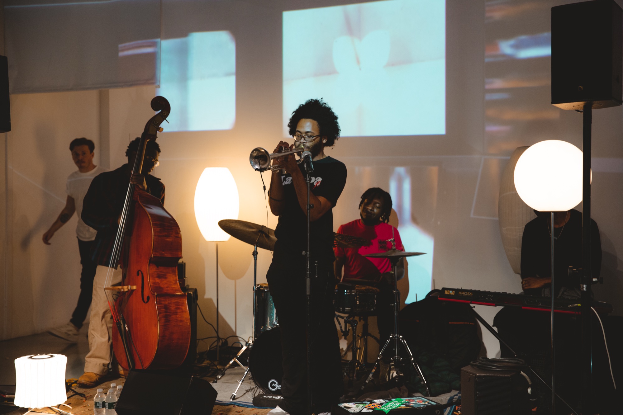 Trumpet player performs with an upright bassist, drummer and keyboardist beside glowing paper lanterns under a projection