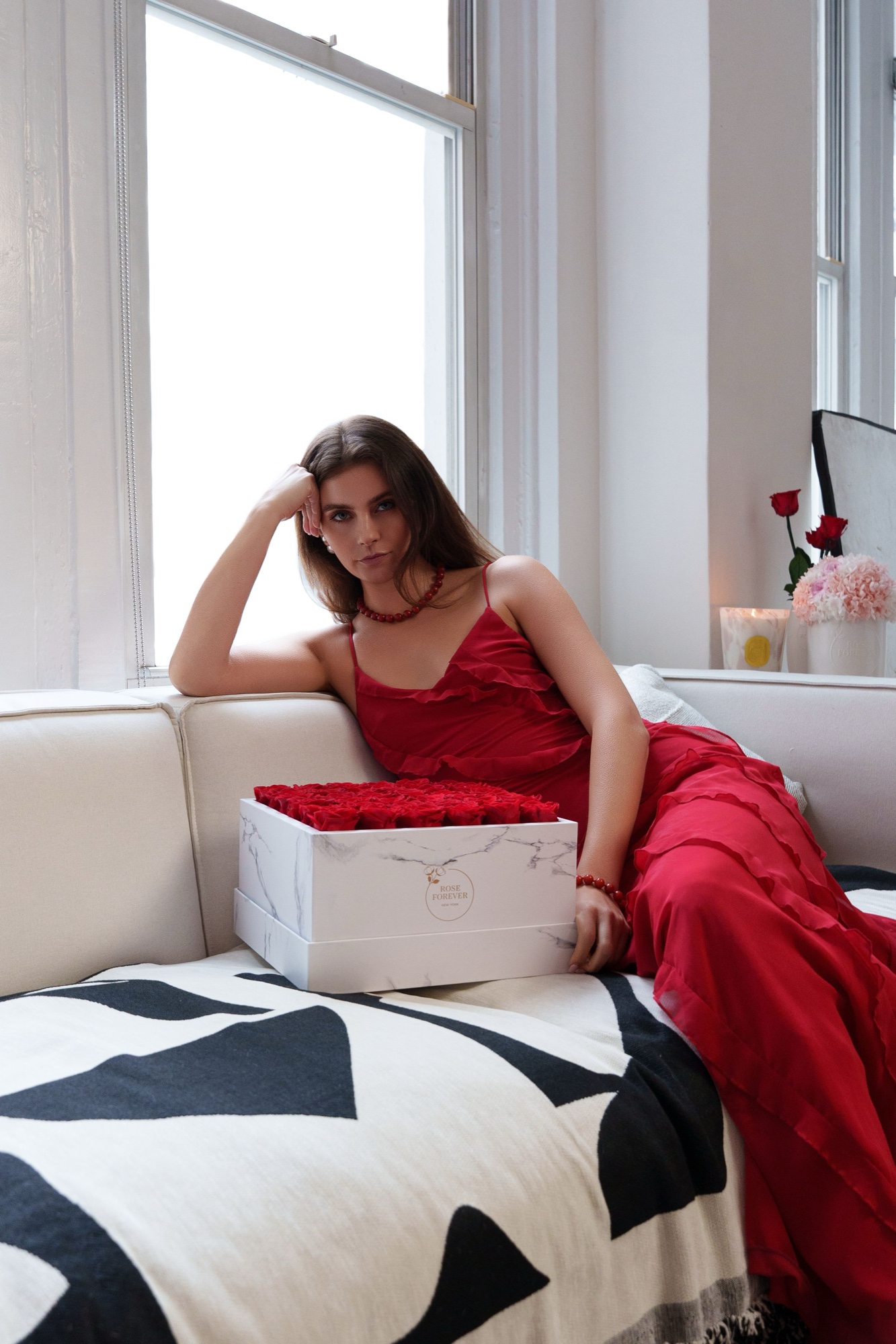 Woman in a red ruffled gown lounges on a sofa with a marble Rose Forever box
