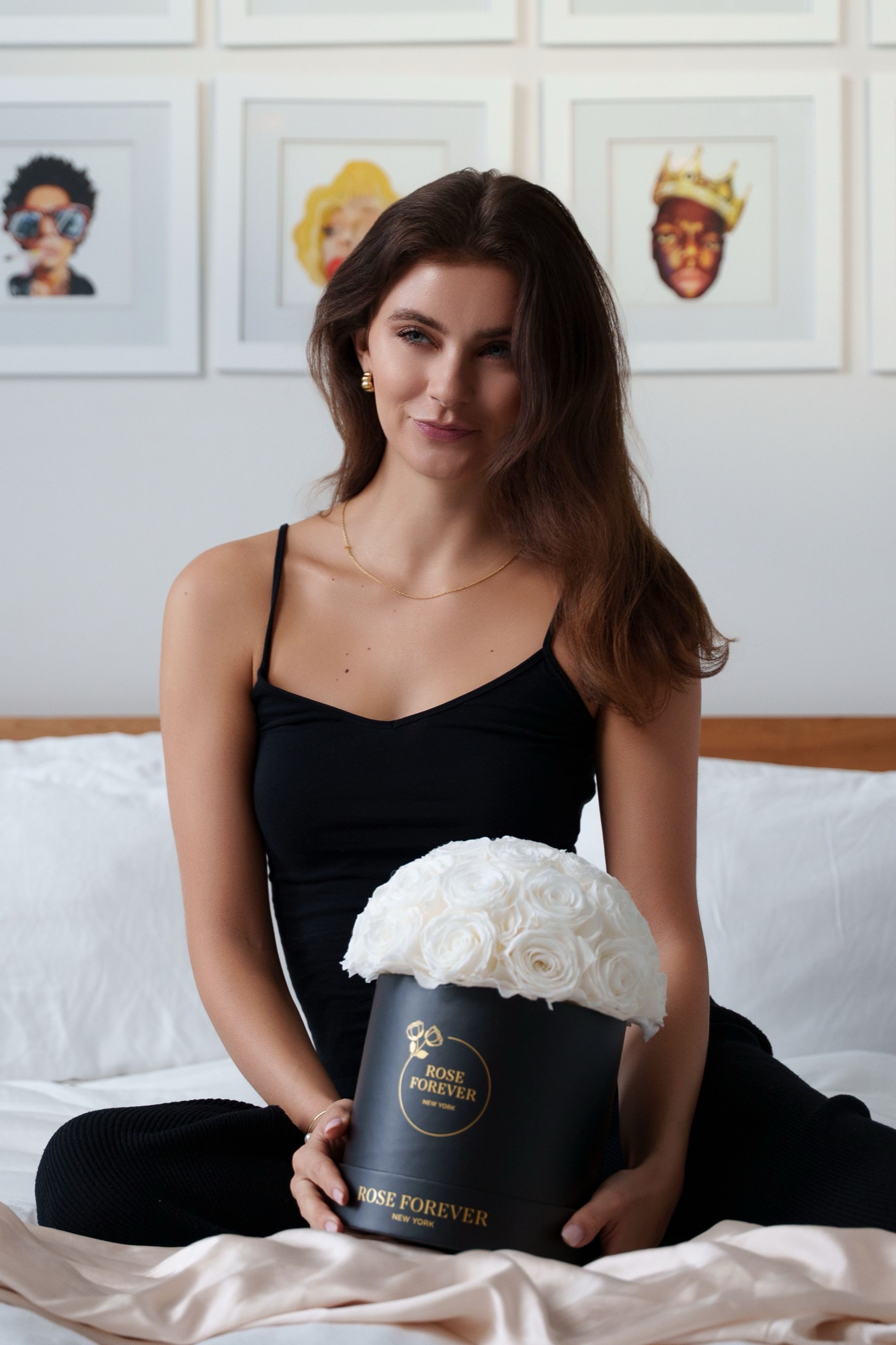 Brunette in black holds a dark Rose Forever box of white roses beneath framed pop portraits