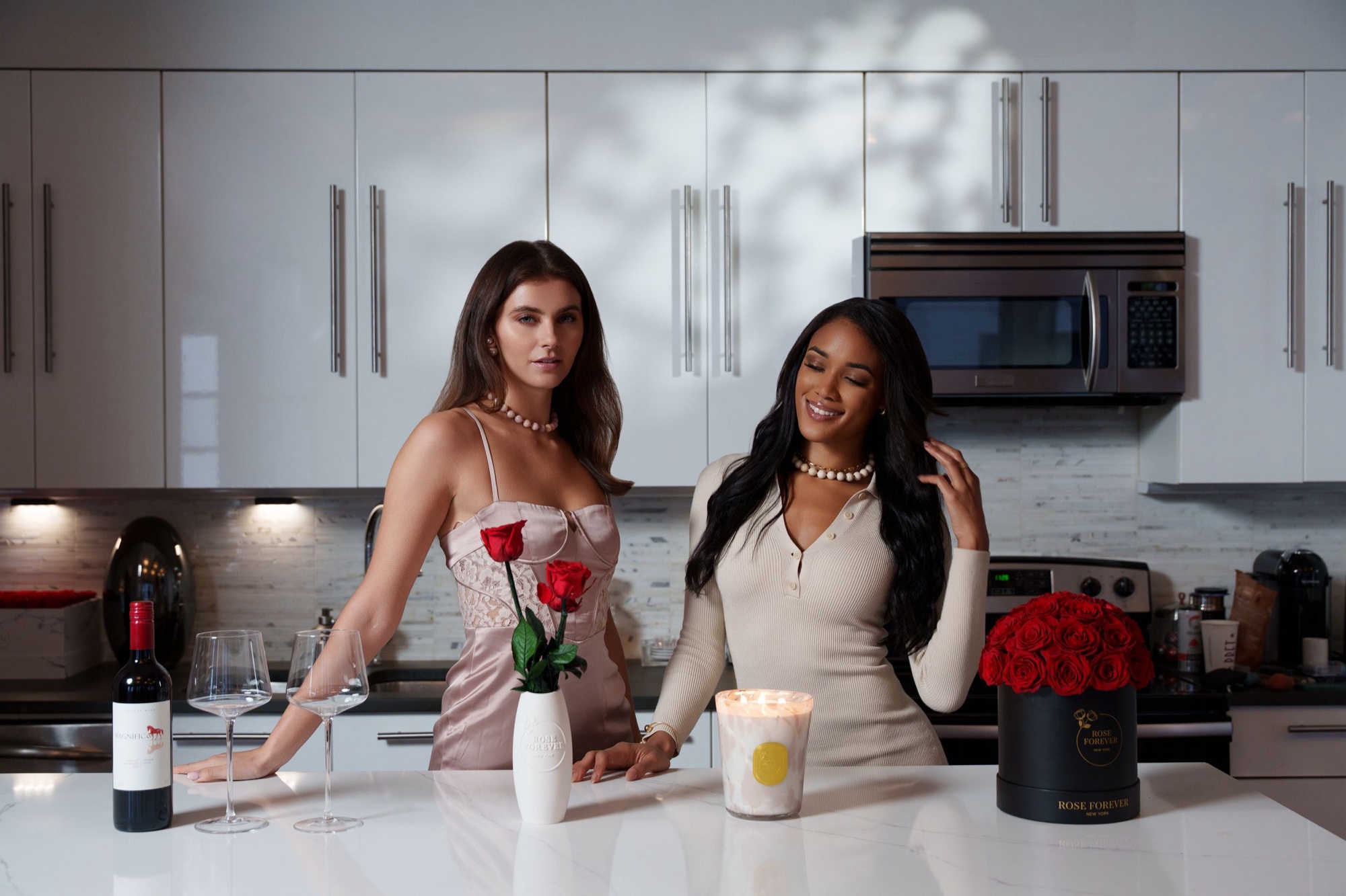 Two models in a white kitchen with wine, candles, and red Rose Forever arrangements