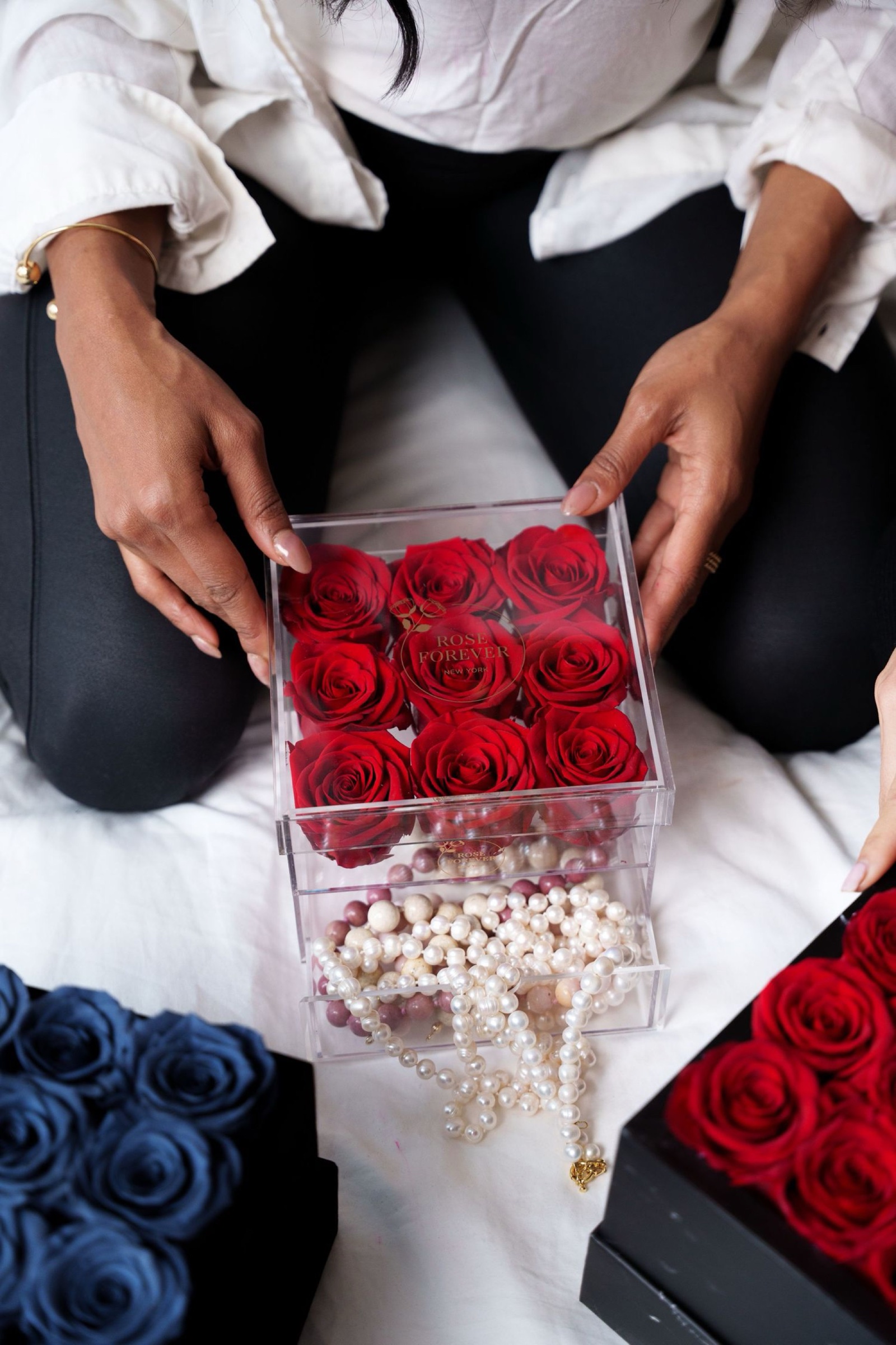A seated woman holds a clear Rose Forever acrylic box of nine red preserved roses, the drawer beneath it open and spilling necklaces made with pearls and stone beads, with navy and red rose boxes on the white bedding around her