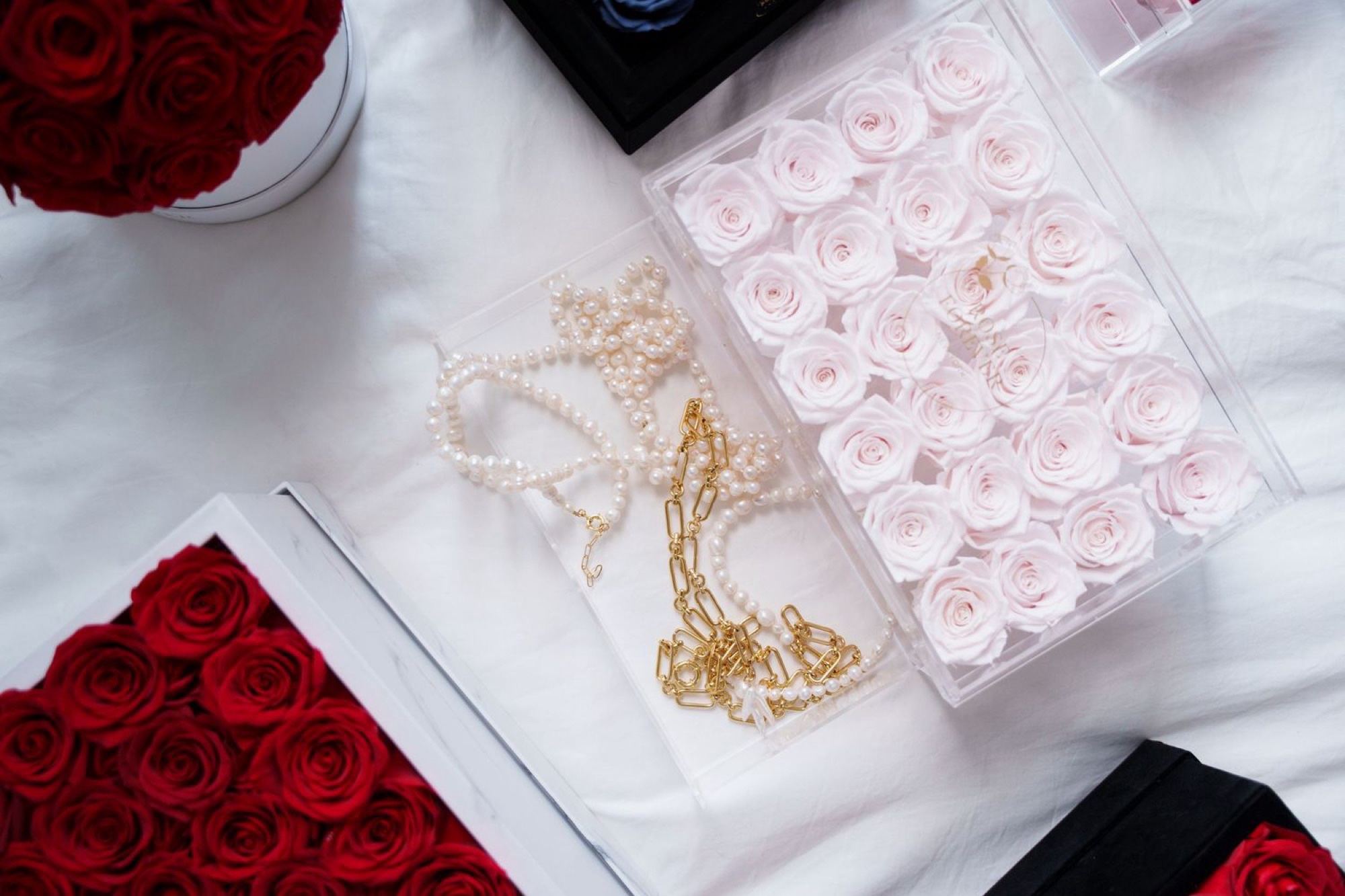 Overhead arrangement of Rose Forever acrylic boxes, pale pink roses in one and red in the others, with pearl strands and a gold chain laid in a clear jewelry tray between them