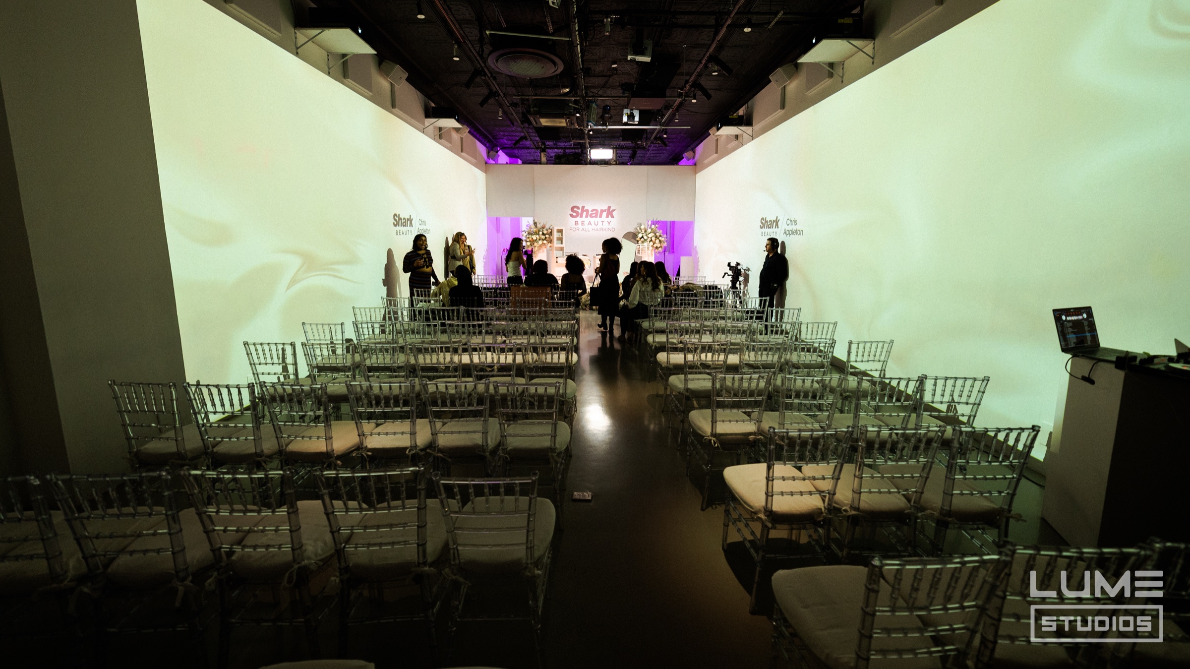 Rows of clear acrylic chairs face a pink and white Shark Beauty stage in a projection lit room
