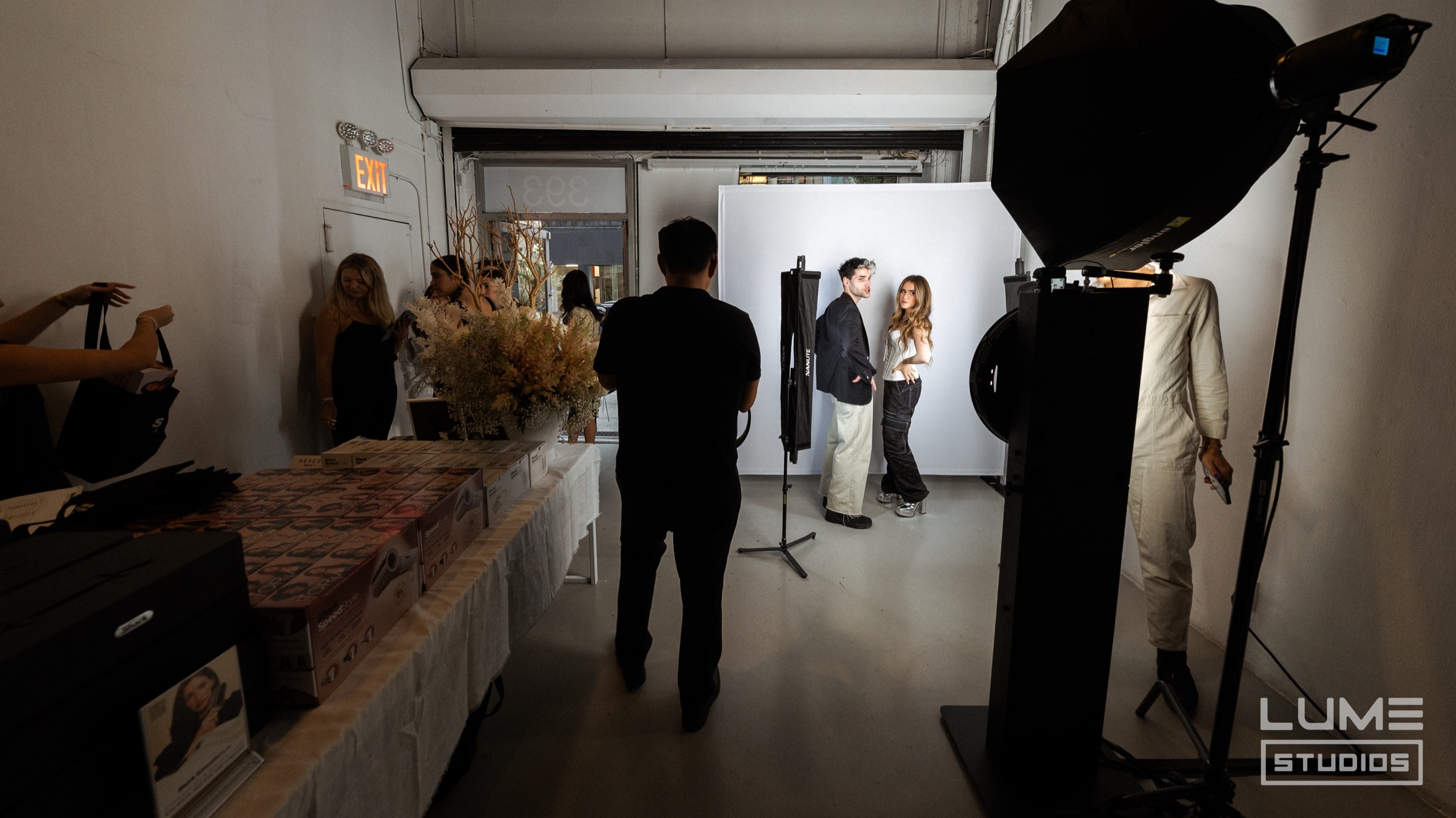 Two guests pose in front of a white backdrop on a photo set while a catering table waits nearby