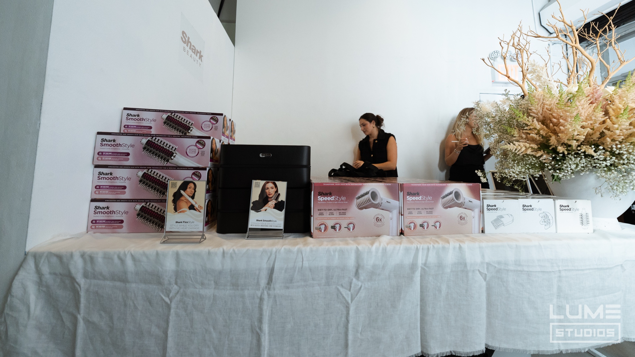 Shark Beauty hair styler boxes stacked on a white draped table beside a tablet display and pale dried flowers