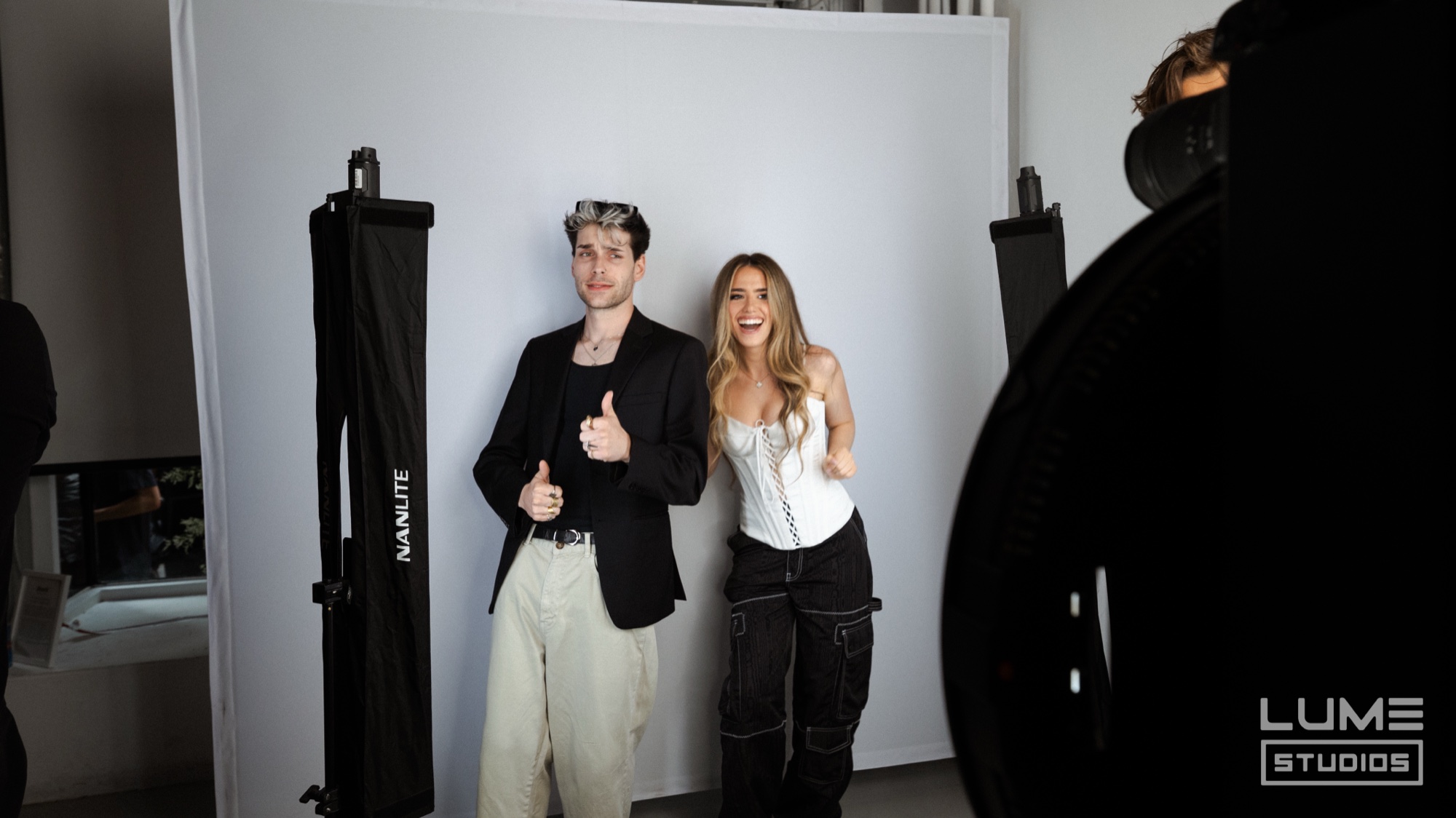 Man in a black blazer and woman in a white corset give thumbs up before a white backdrop and softboxes