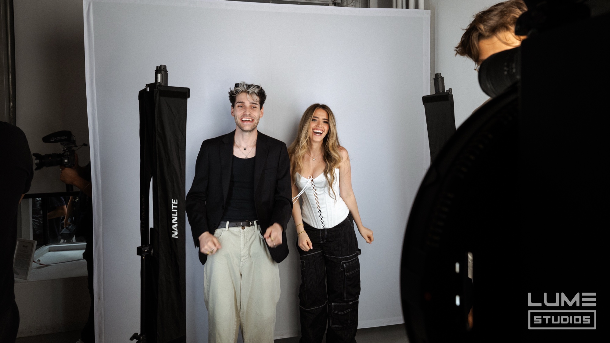 Smiling pair pose on a white backdrop between softbox lights in a black blazer and white corset