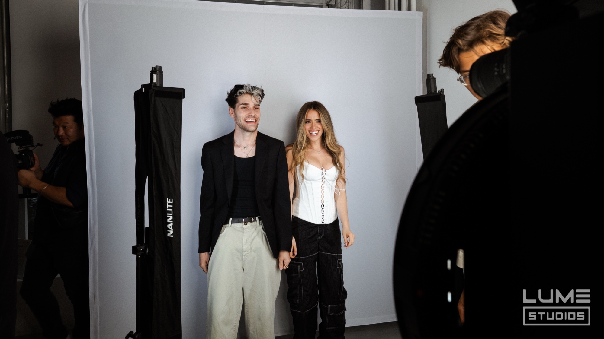 Two people pose on a white backdrop between softbox lights while a video crew films from the sides