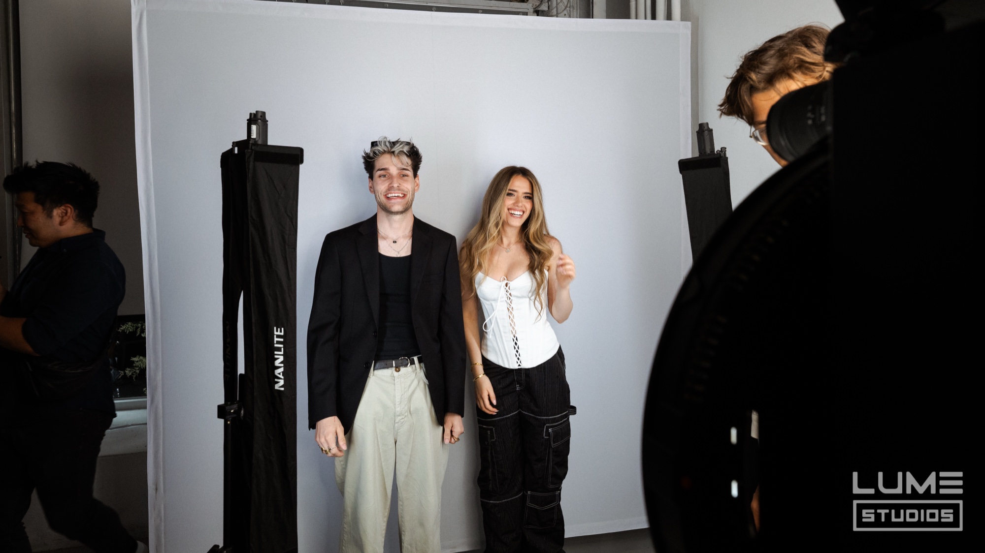 Man in a black blazer and woman in a white corset pose smiling against a white backdrop flanked by softboxes