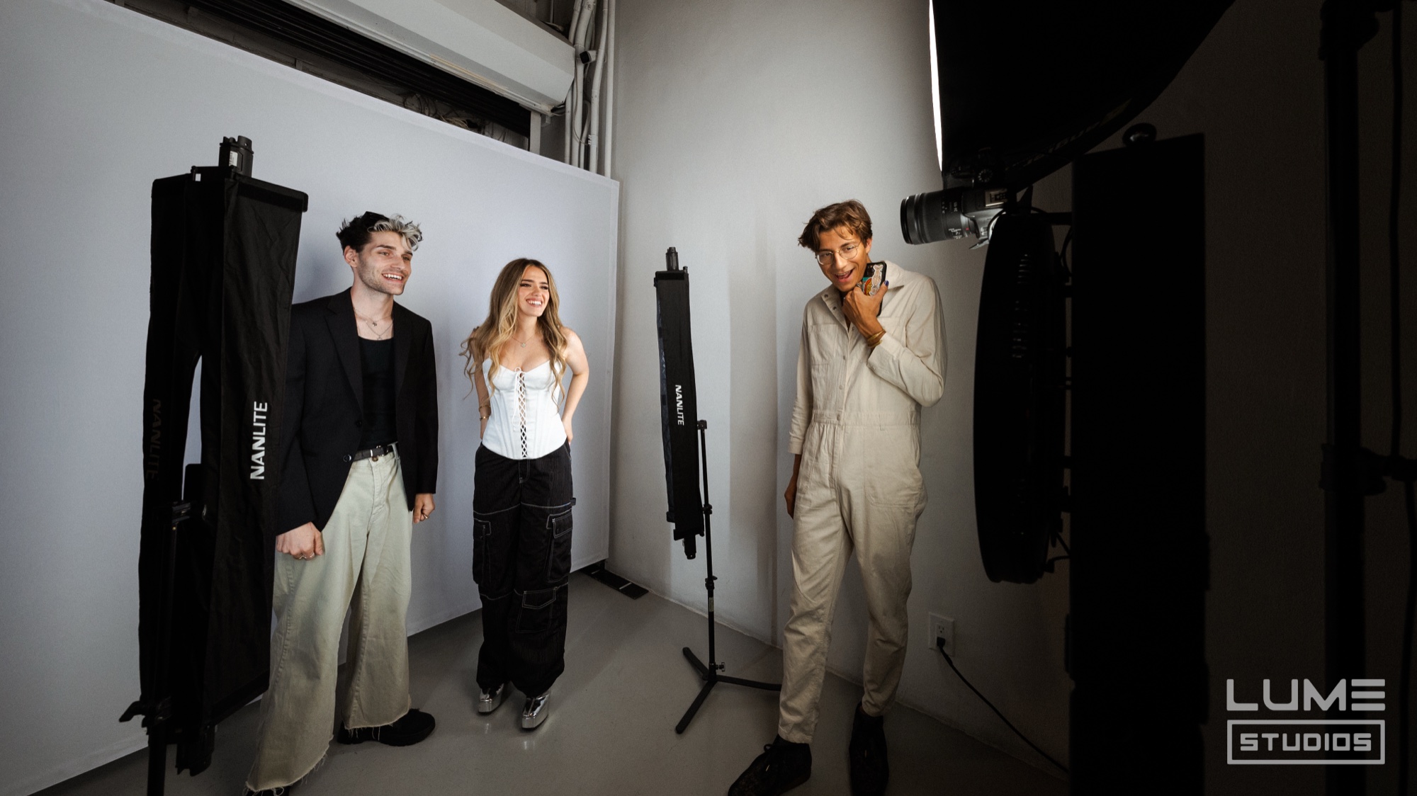 Three people on a grey studio backdrop, one in a cream jumpsuit laughing into his hand