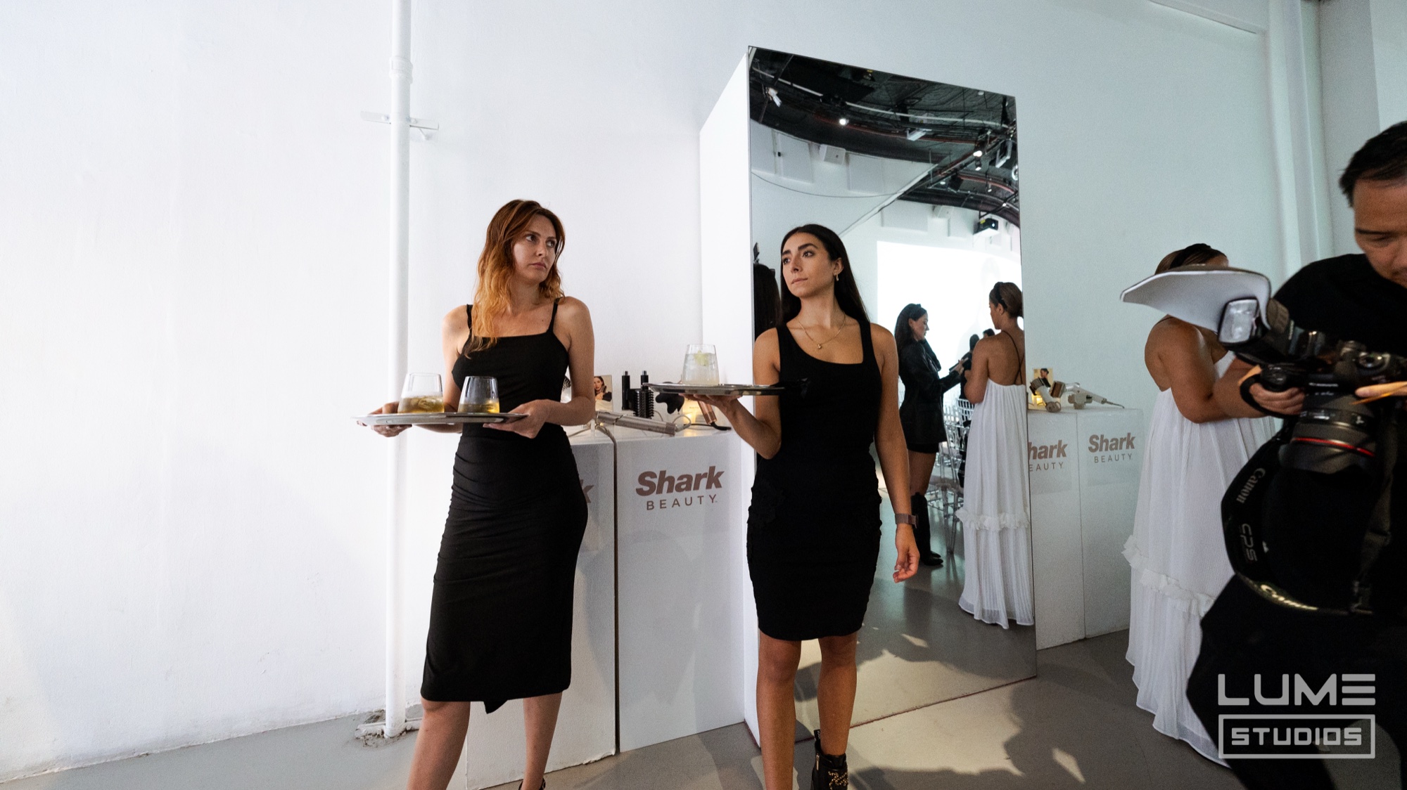Two servers in black dresses carry trays past a Shark Beauty sign while a photographer shoots nearby
