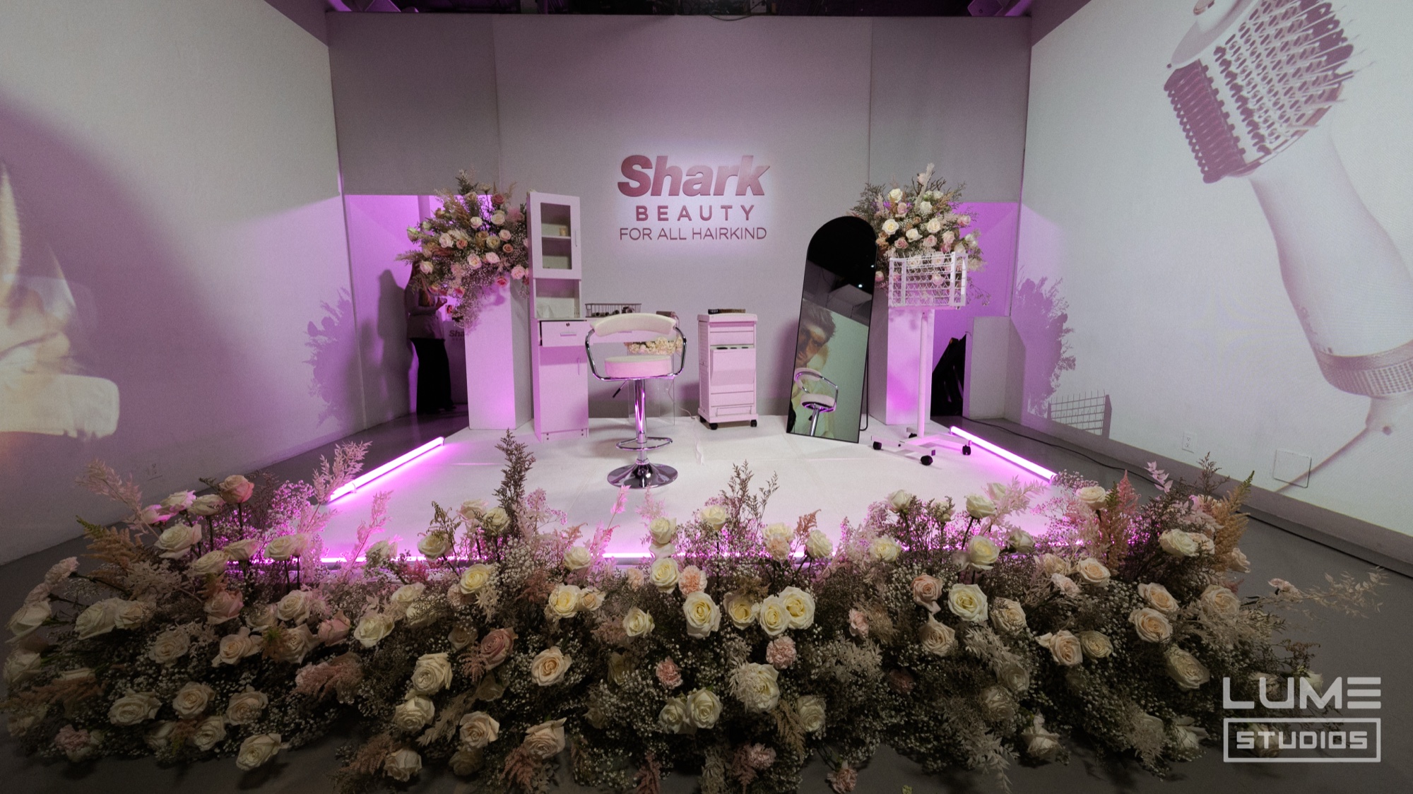 Pink lit Shark Beauty stage with a salon chair, white flower arrangements and a hairdryer projected on the wall