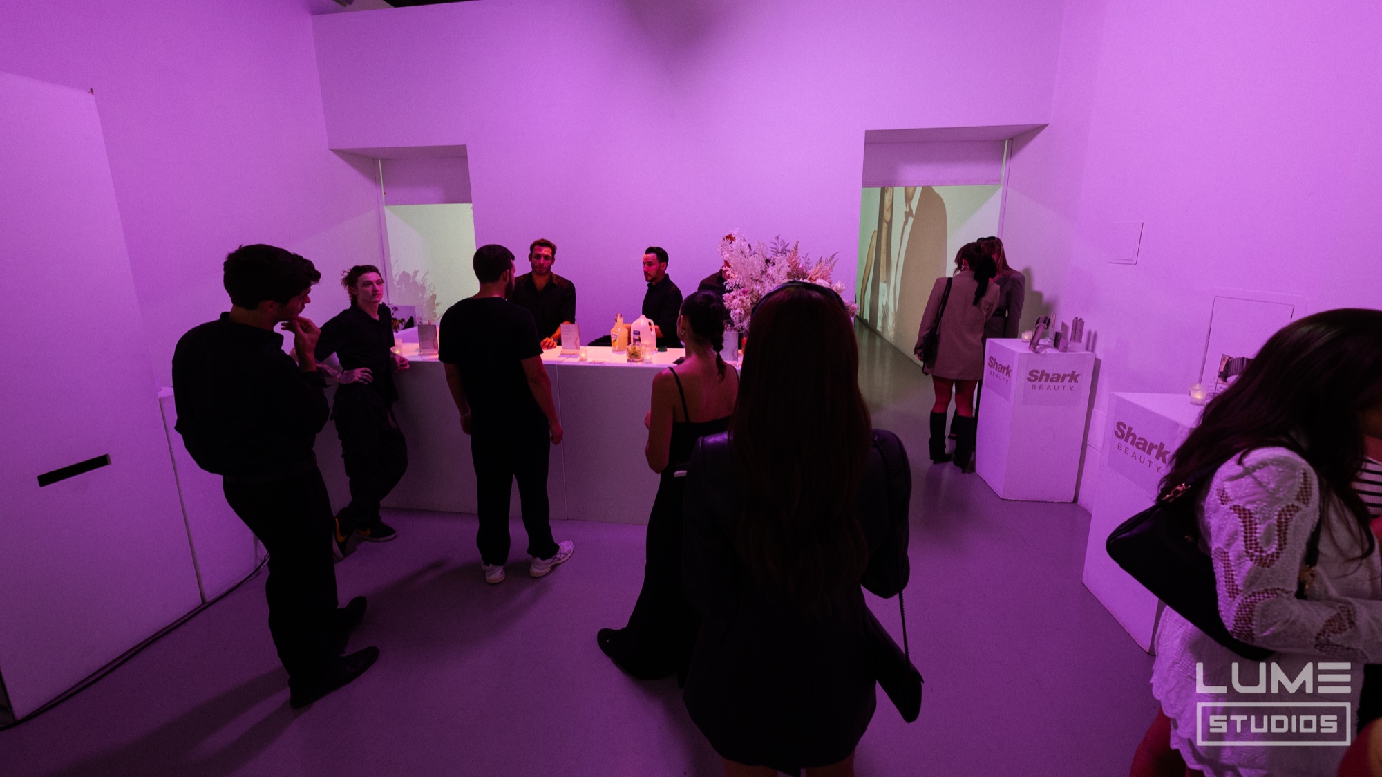 Guests in black mingle around a white counter in a room washed in purple light