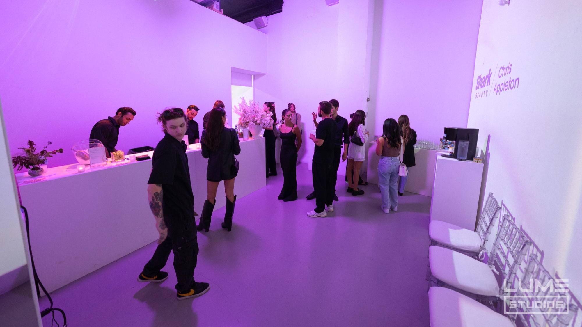 Guests mingle beside a white bar in a lavender lit room with Shark Beauty x Chris Appleton wall signage