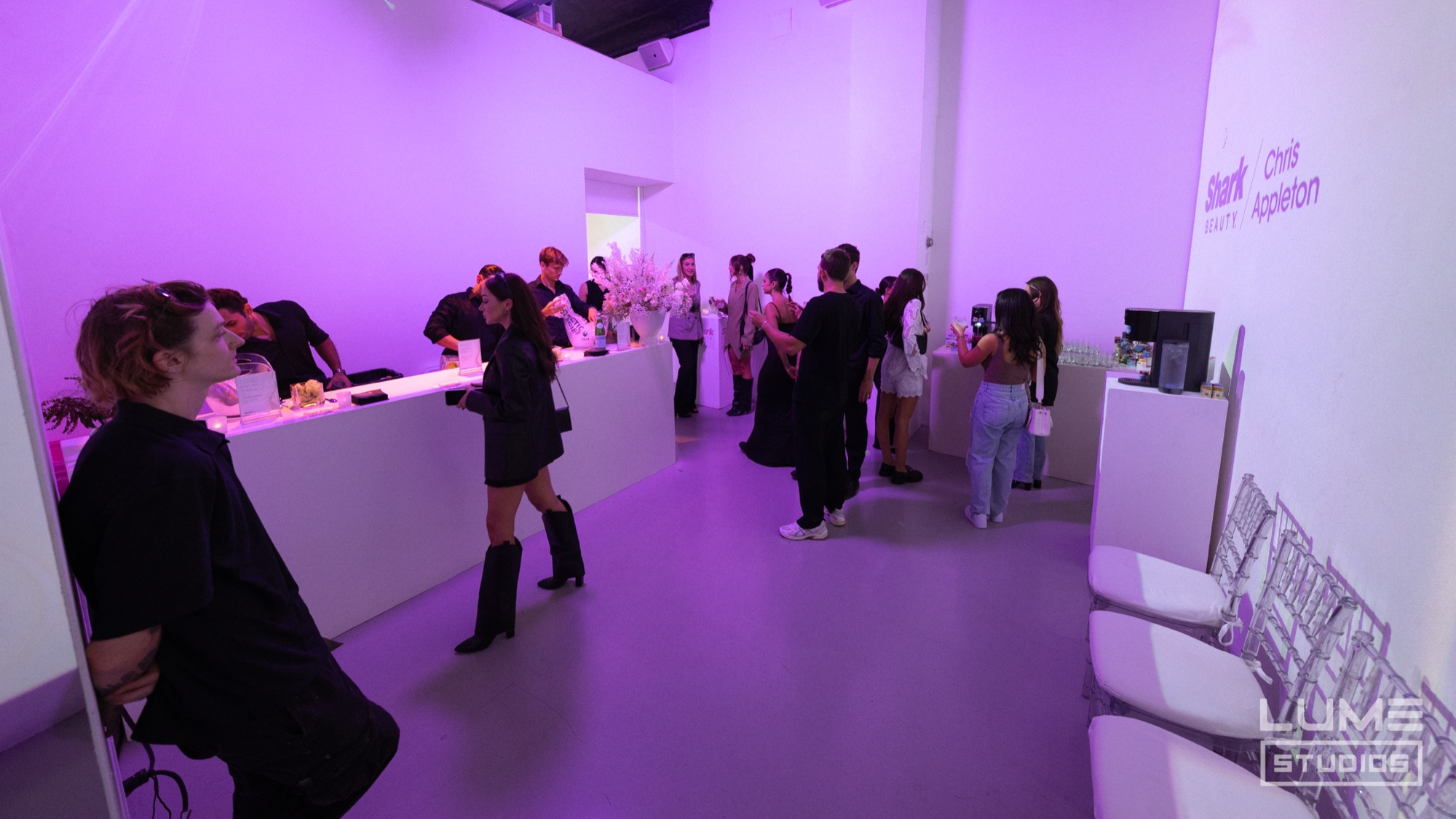 Guests gather around a white bar in a purple lit room with Shark Beauty signage on the wall