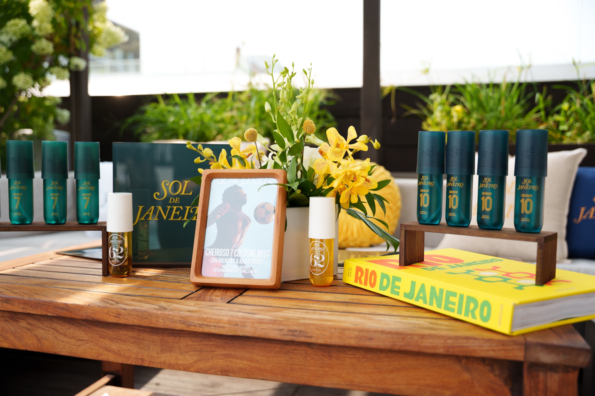 Green Sol de Janeiro spray bottles, yellow flowers and a Rio de Janeiro book arranged on a wooden table outdoors