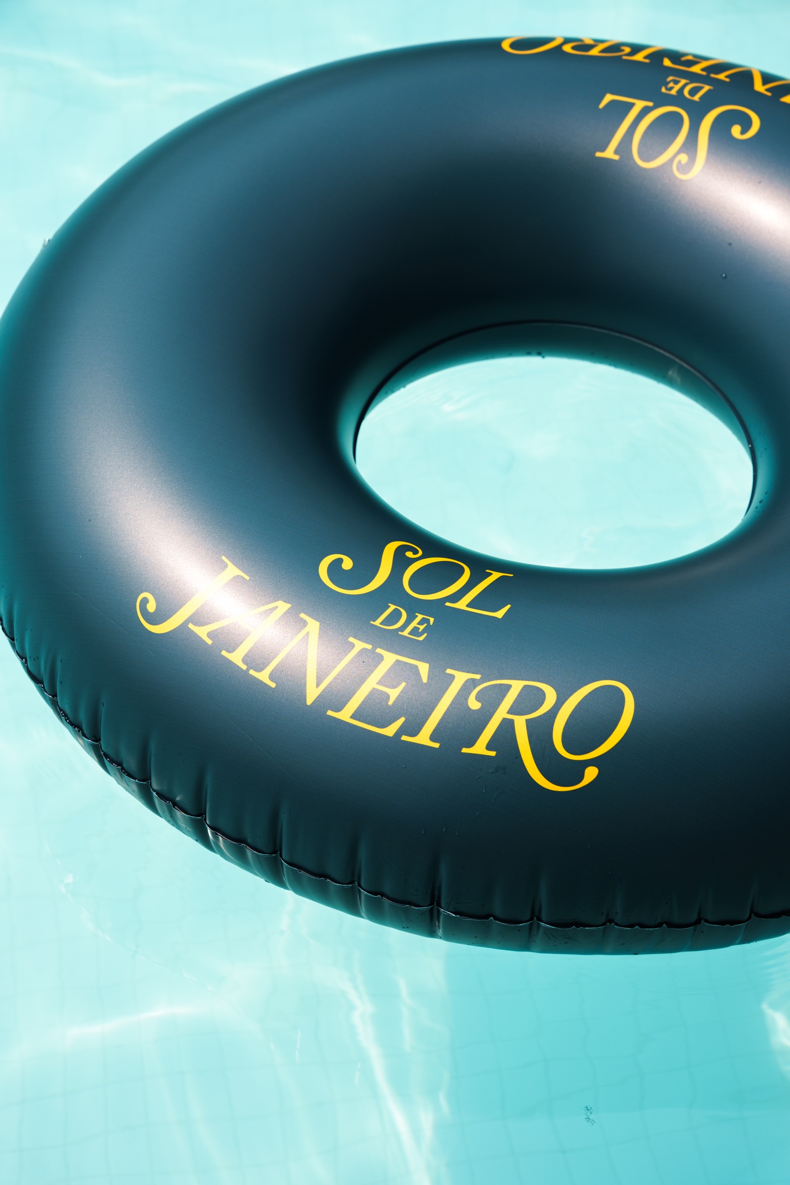 Dark green inflatable ring printed with Sol de Janeiro in yellow floating on clear turquoise pool water