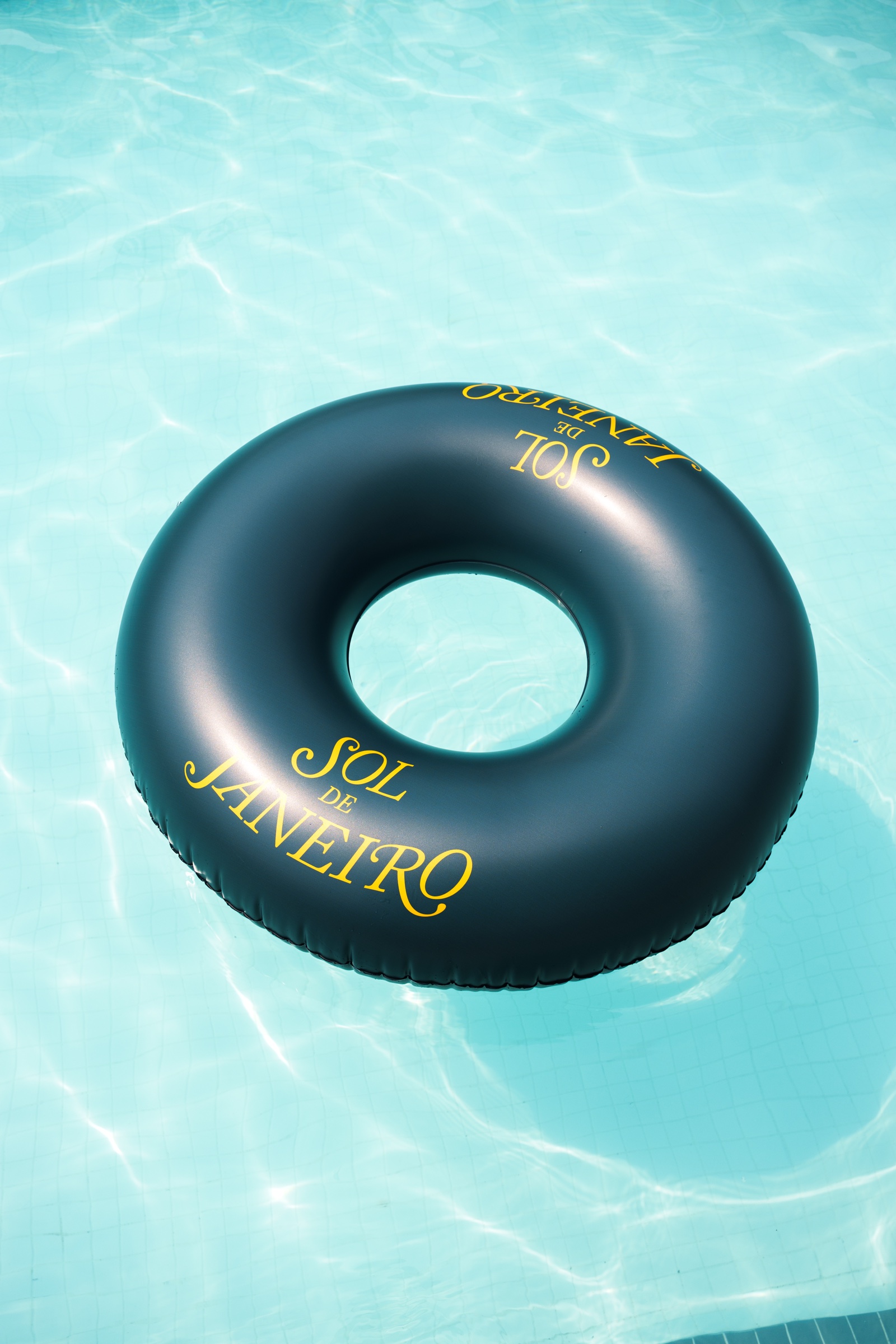 A black inflatable ring printed with Sol de Janeiro in yellow floats on clear turquoise pool water