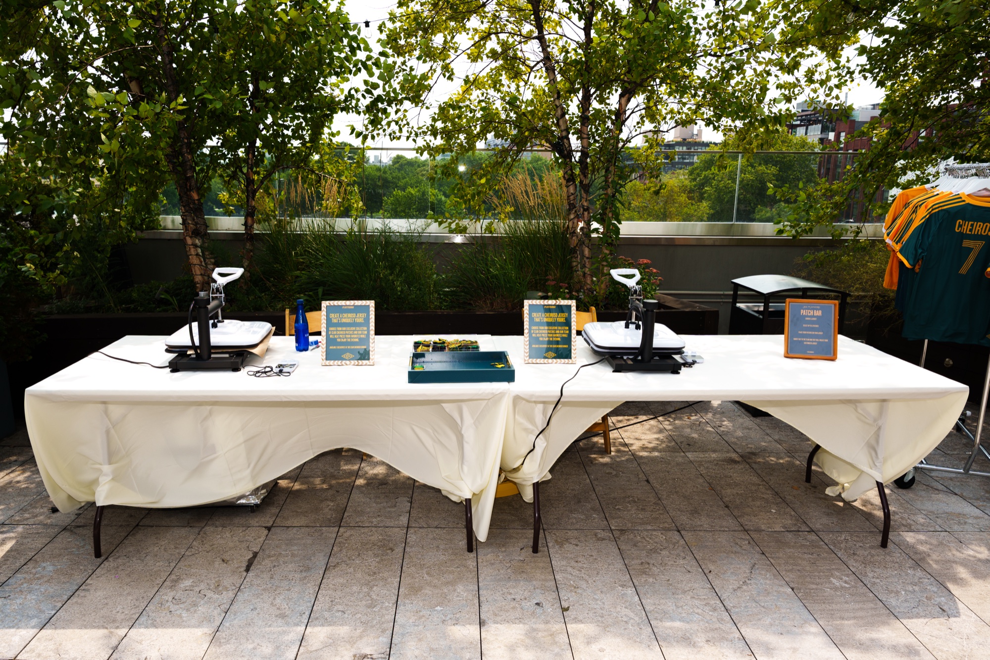 Two heat presses on a white draped table beside a rack of yellow tees on a leafy rooftop