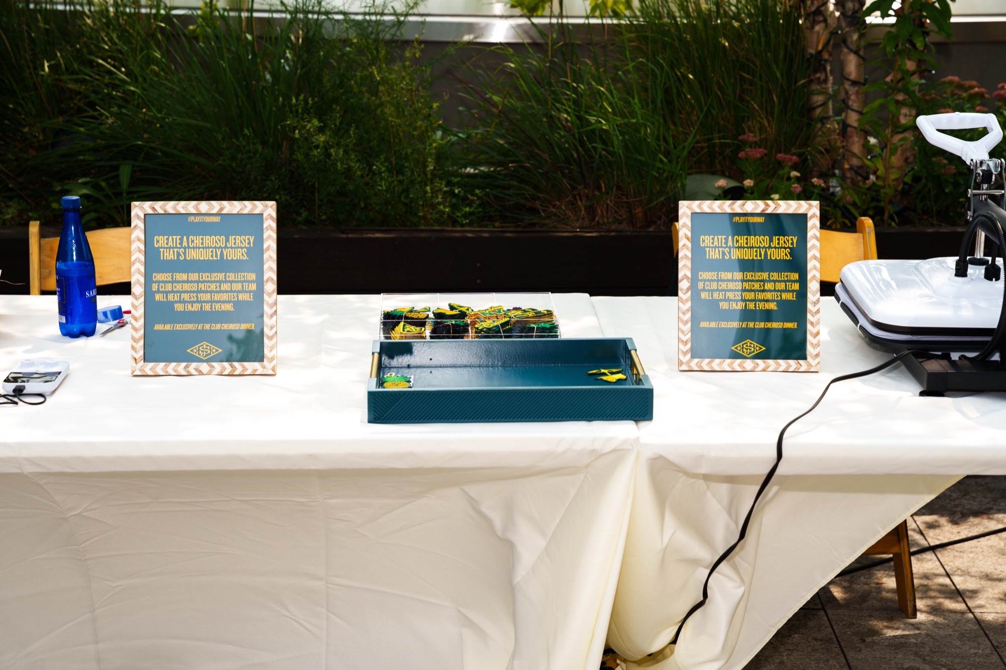 A white draped table holds a tray of green and yellow patches, two printed signs and a heat press