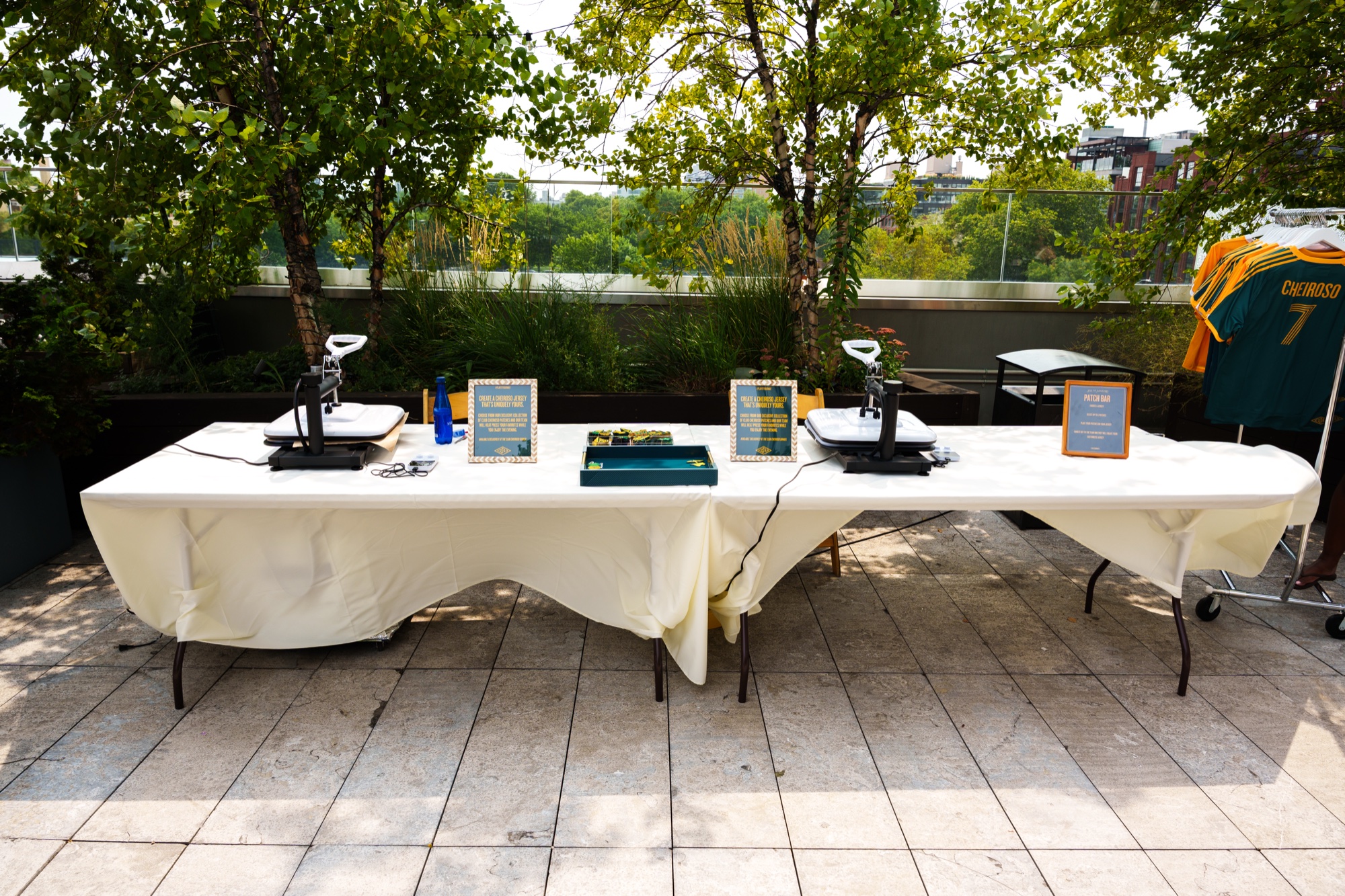 Two heat press stations and small signs set on a white draped table on a rooftop terrace with trees behind