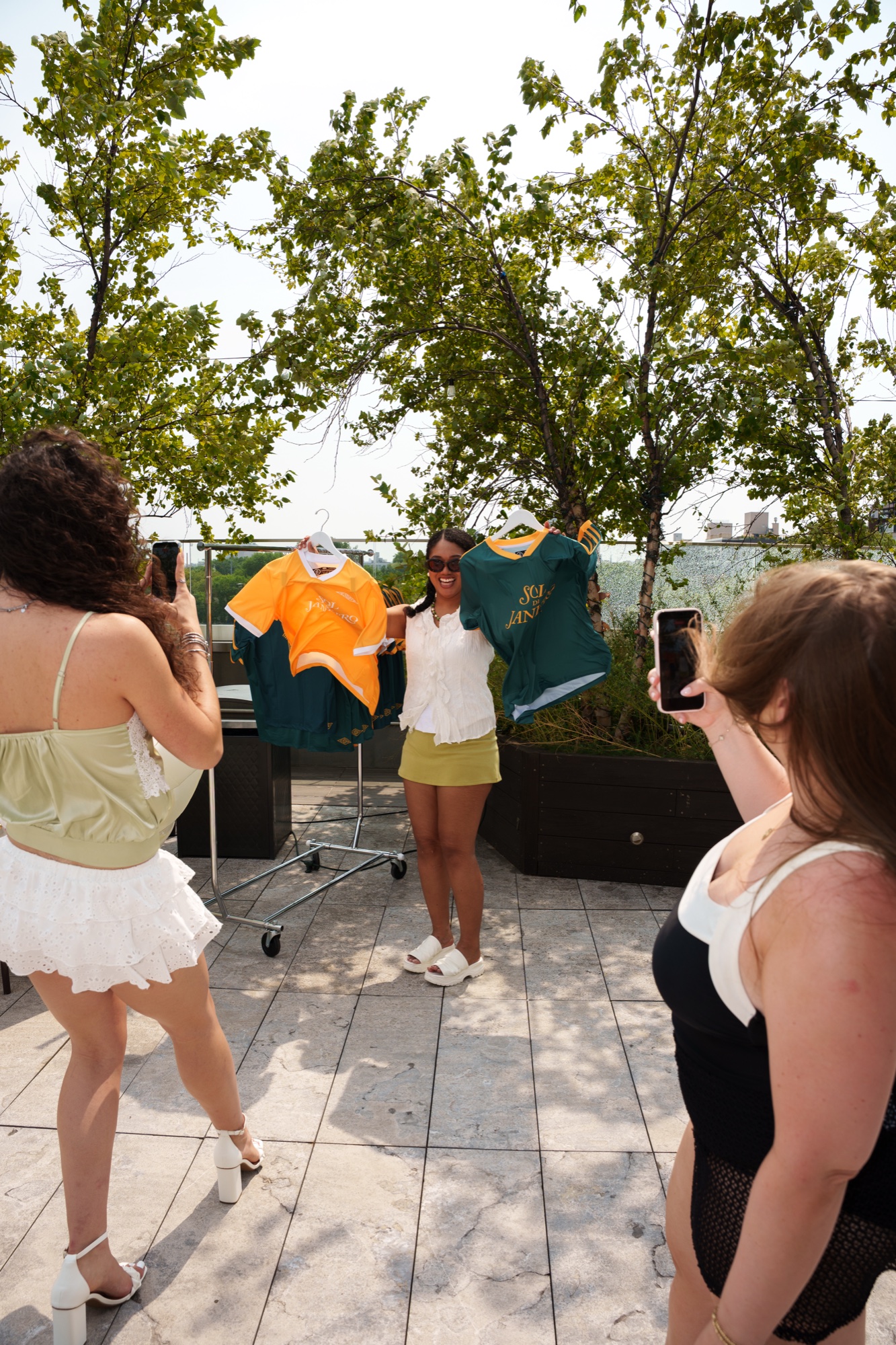 A woman holds up orange and green jerseys on a sunny rooftop while two friends photograph her with phones