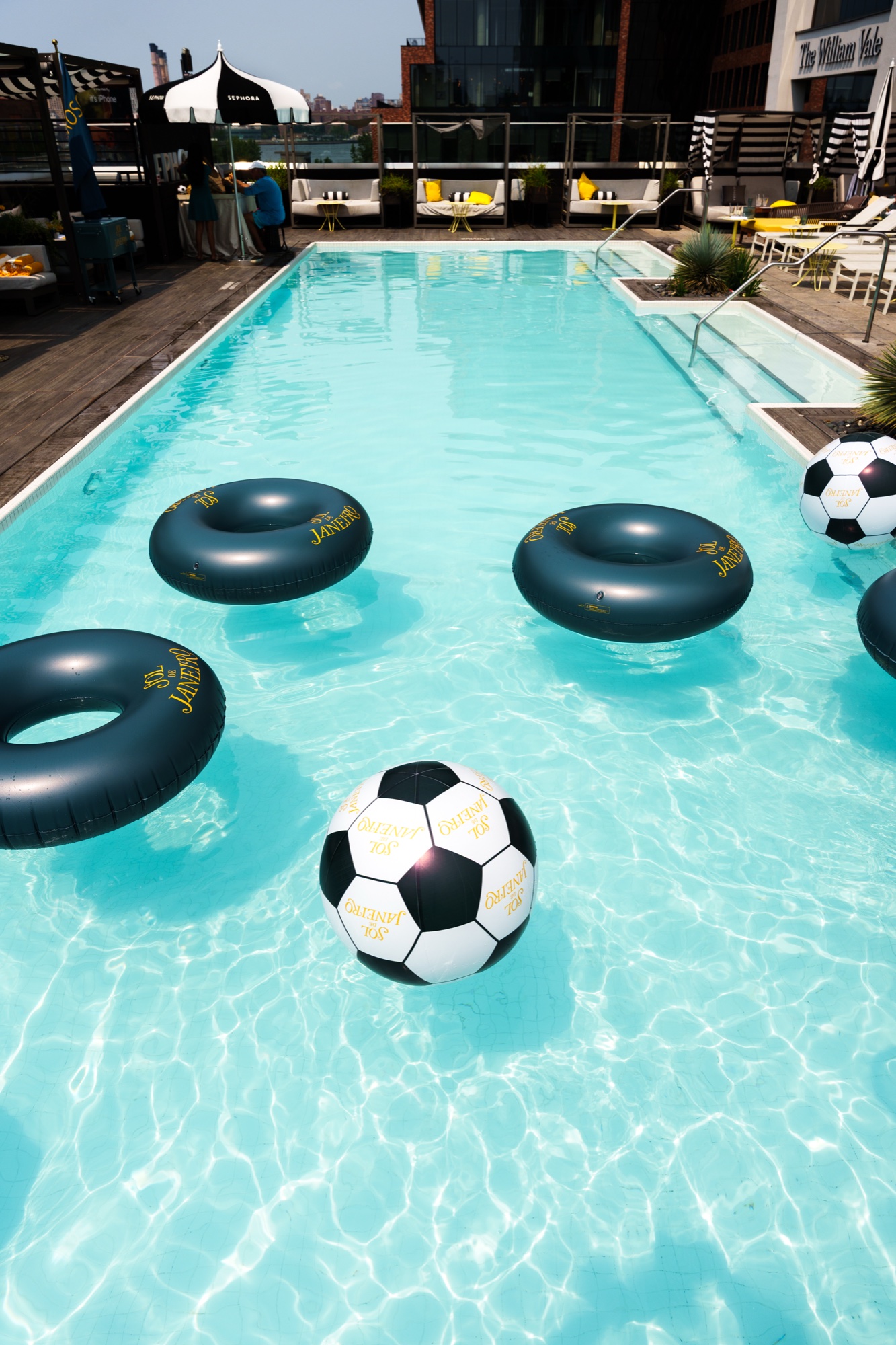 Black inflatable rings and soccer balls float in a rooftop pool beneath The William Vale sign
